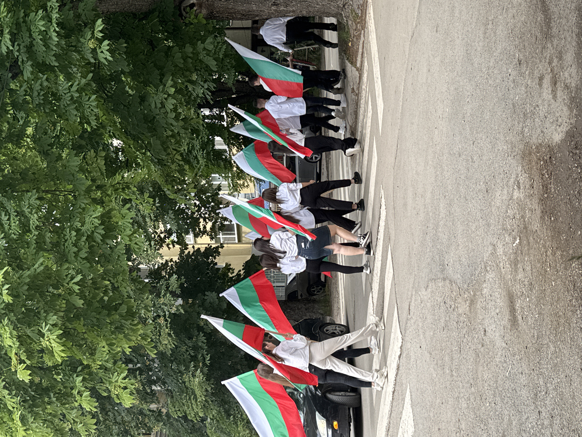The image displays a group of young people, likely students, participating in a procession or parade in Targovishte, Bulgaria, during daylight hours. They are captured walking in a line across a pedestrian crosswalk on a paved street.
Each person in the foreground and many in the background are holding a Bulgarian flag, which features horizontal stripes of white, green, and red. The participants are largely dressed in coordinated attire, consisting of white long-sleeved tops, such as shirts or hoodies, paired with dark trousers or skirts, and often sneakers. Their faces are mostly obscured or turned away from the camera.
The scene is set on a street bordered by lush green trees with dense foliage, suggesting a verdant urban environment. In the background, glimpses of buildings can be seen through the trees, alongside parked cars on the side of the road. The lighting indicates it is daytime, possibly a bright but overcast day, as no strong shadows are visible. The event appears to be a public display of national pride or a commemoration, consistent with a national holiday or cultural celebration in Bulgaria. No discernible text is visible in the image.