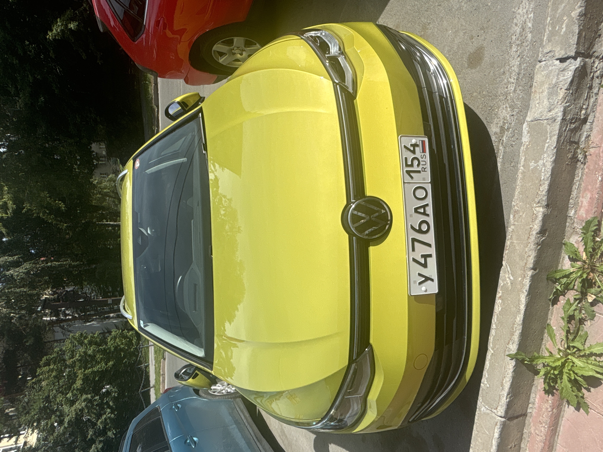 A bright yellow Volkswagen Golf is parked on a street in Novosibirsk, Russia, as evidenced by the license plate which reads "y476AO 154 RUS". The car is facing the camera directly and is positioned between a blue car on its left and a red car on its right. The street appears to be in a residential area, with trees and buildings visible in the background. The sun is shining, casting shadows on the ground. The car's headlights are on, and the license plate is clearly visible. The car appears to be in good condition.