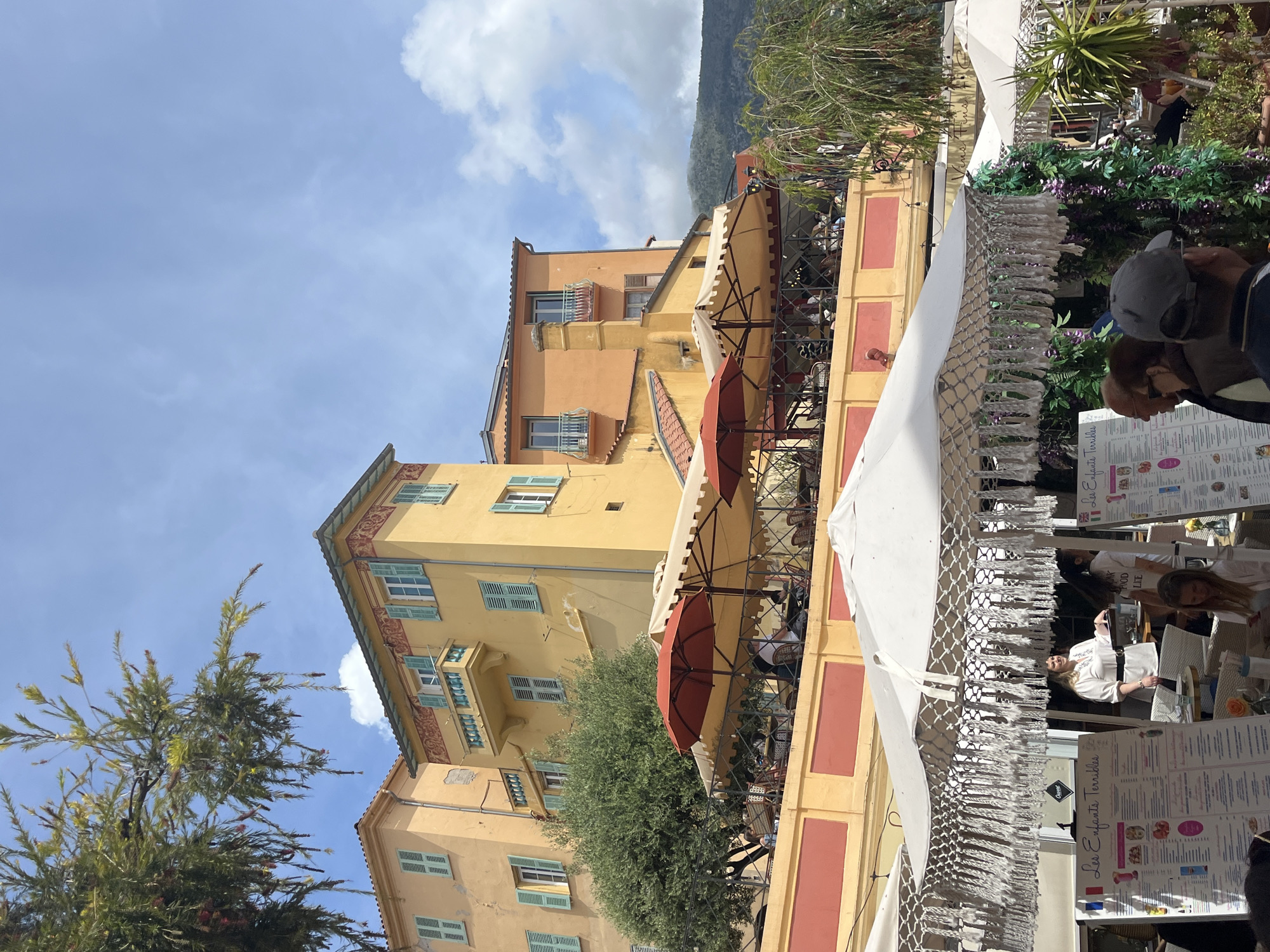 The image captures a street scene in Menton, France, characterized by its colorful architecture and outdoor dining.
**Environment:** A sunny day with a blue sky and scattered clouds. Buildings are painted in shades of yellow and orange, with decorative details around windows and cornices. A mountain range is visible in the background, adding depth to the scene.
**Objects:** A prominent feature is the outdoor restaurant seating area. Red and white umbrellas provide shade for the diners. Tables and chairs are visible, suggesting an active dining environment. Tassels are at the edge of the canopy. Menu boards are displayed.
**People:** Patrons are seated at the outdoor tables, engaged in dining and conversation. Waitstaff are working. Pedestrians pass by.