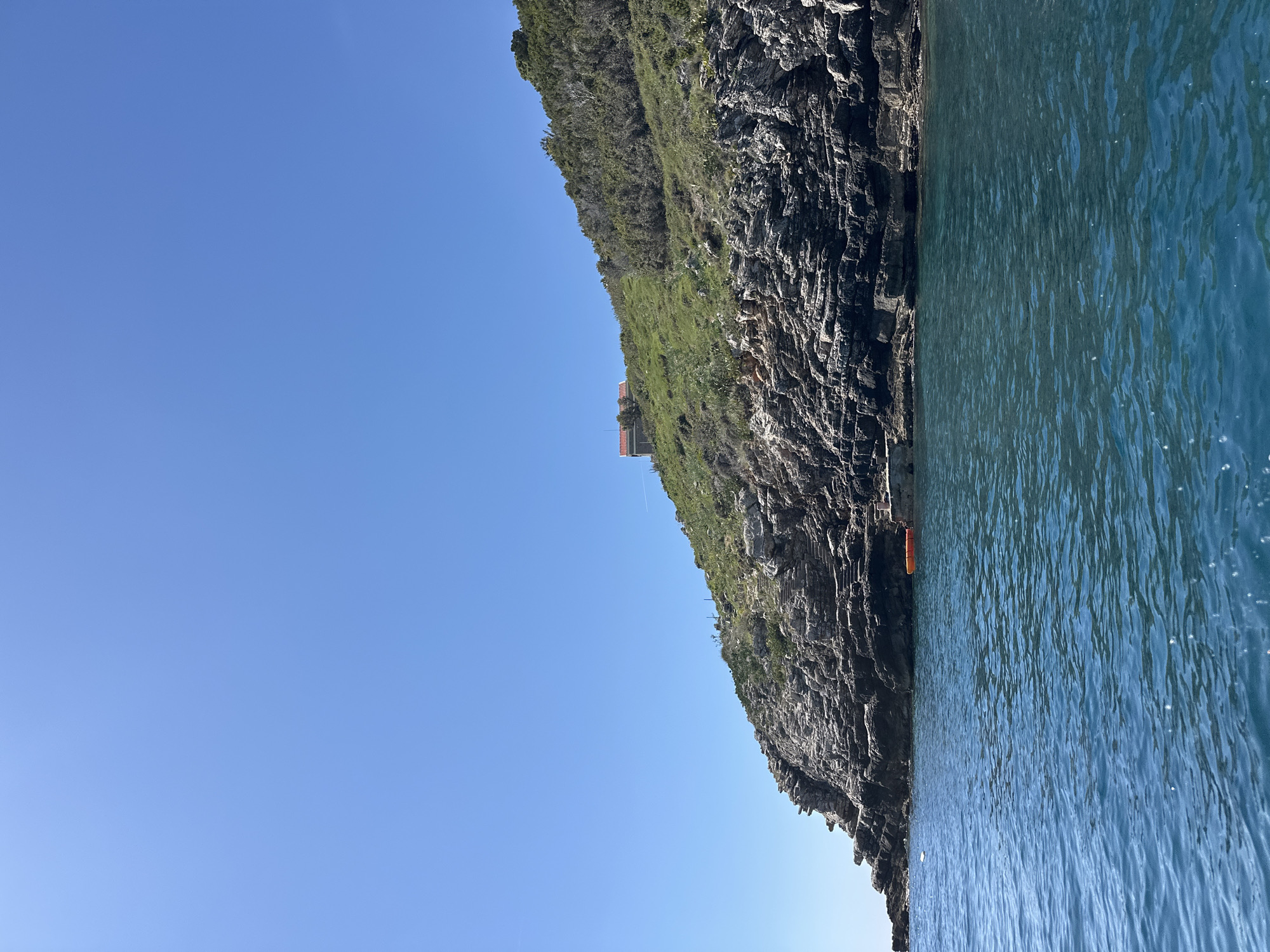The image depicts a cliff face in Budva, Montenegro, meeting the Adriatic Sea under a clear, blue sky. The cliff is composed of dark, layered rock, with patches of green vegetation clinging to its upper reaches. An orange kayak is visible along the base of the cliff, close to the water's edge. A building with an orange roof can be spotted on the clifftop. The water is a deep turquoise, reflecting sunlight.