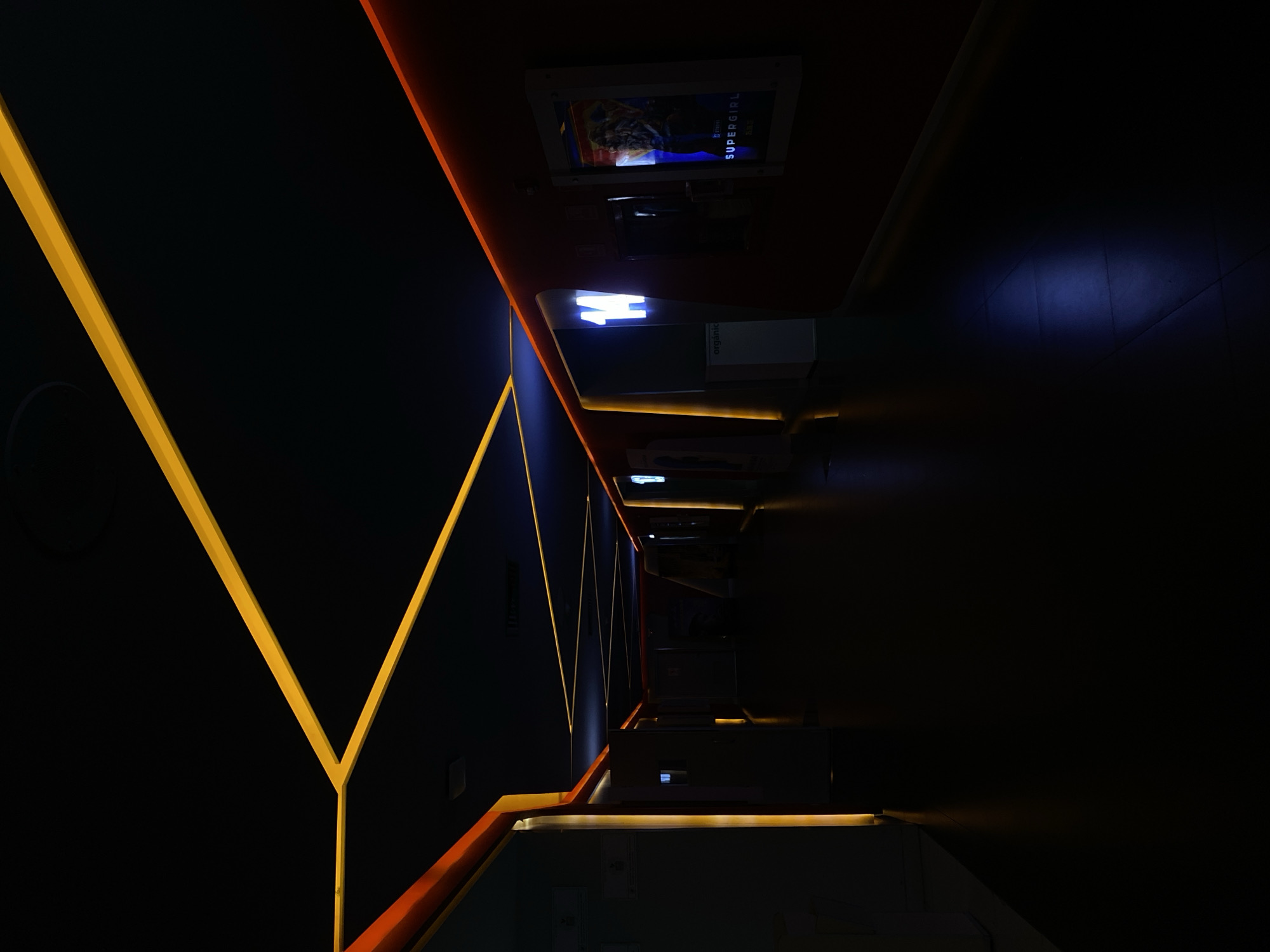 This media file captures a dark, modern interior space, likely a corridor or hallway, from a highly tilted perspective. The scene is dominated by striking linear light strips in vibrant yellow and orange, which delineate the architectural structure against a deep black background. These glowing lines run along what appear to be the walls and ceiling, forming sharp geometric patterns that extend into the distance, creating a strong sense of depth.
On the right side of the image, which would be the upper wall or ceiling if viewed upright, several illuminated display screens are visible. One screen prominently displays the word "SUPERGIRL" in white text, accompanied by an image of a character. Above it, another screen is lit but its content is less clear. Further along the corridor, a bright, stylized white letter "H" is visible on an illuminated sign, and below it, the word "organic" can be faintly discerned.
The space is entirely devoid of people, drawing focus to the deliberate architectural design and artificial lighting. The dark environment, accentuated by the bright, contrasting light strips and digital displays, creates a futuristic and somewhat mysterious atmosphere. The yellow lights form angular, converging lines on one side, while the orange lights provide a continuous border on the other. The low-light setting emphasizes the luminescence of the light elements, making them stand out dramatically. This aesthetic suggests a commercial or public venue, such as a cinema, entertainment complex, or exhibition hall.