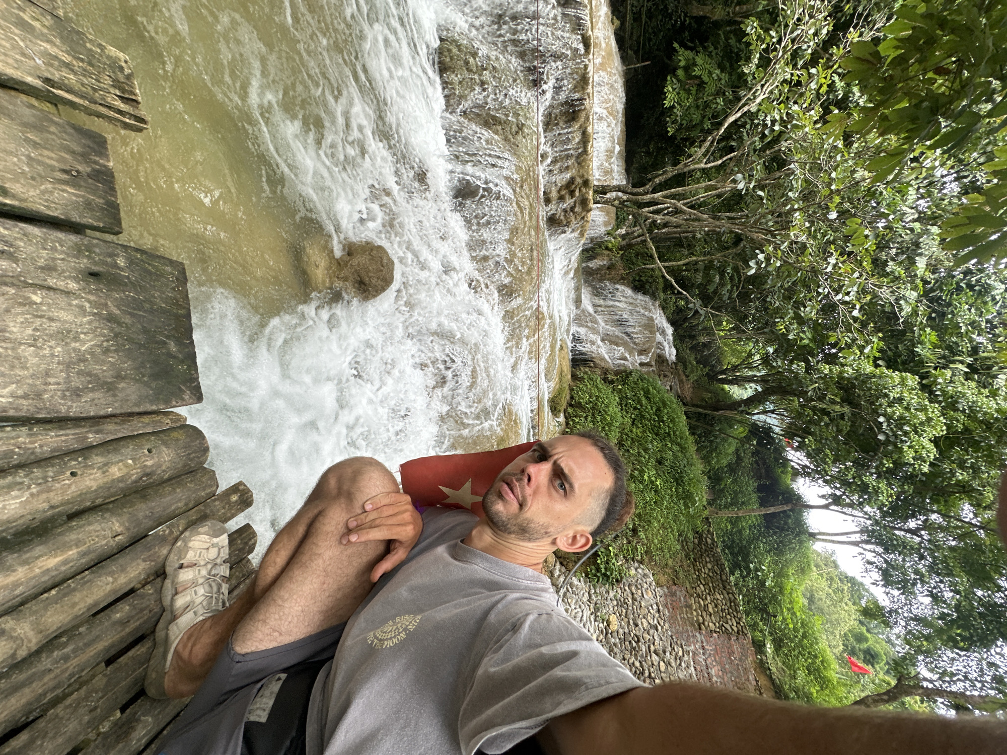 In the foreground, a man crouches on a wooden walkway made of rough planks. He is wearing a grey t-shirt and dark shorts, with light-colored sandals. A red flag with a yellow star is tucked into his waistband. The man is looking directly at the camera with a neutral expression. Behind him, a multi-tiered waterfall cascades down rocky formations into a wide, turbulent river. The surrounding environment is lush and green, with dense trees and vegetation covering the hillsides. The location appears to be a natural landscape, possibly a tourist attraction in the Cổ Lũng Commune, Vietnam, given the presence of the Vietnamese flag. The water in the river is a cloudy brown color, and the spray from the waterfall creates a misty atmosphere. The wooden walkway extends across the bottom of the frame, providing a vantage point over the rushing water.
