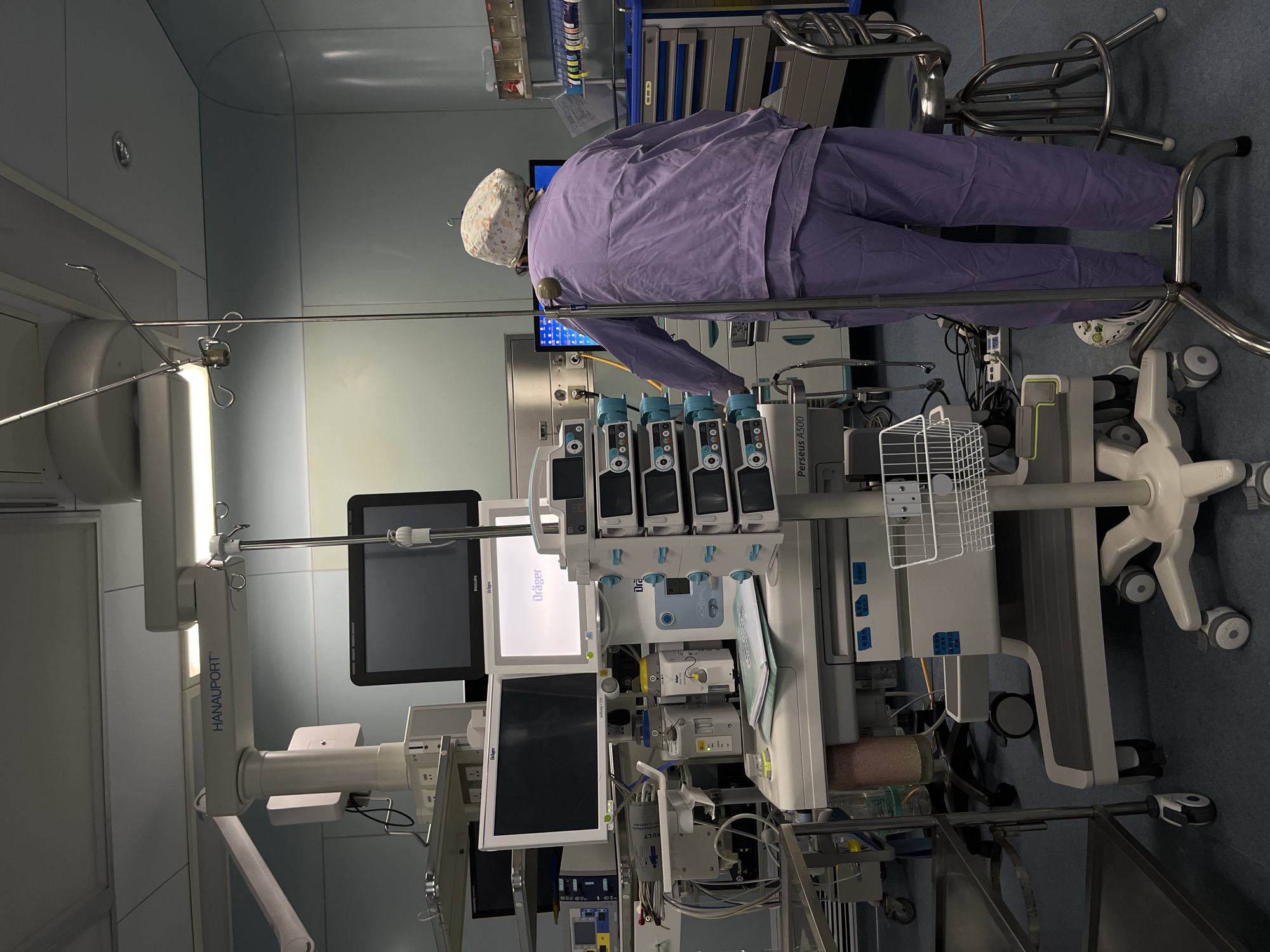 The image captures a sterile medical environment, likely an operating room or intensive care unit, in Yuzhong District, China. A single medical professional, viewed from behind, is the primary subject. They are dressed in purple scrubs and a patterned surgical cap, standing and appearing to interact with or monitor a sophisticated array of medical equipment.
The room is densely packed with advanced medical devices. To the left, a prominent, ceiling-mounted medical supply unit or boom system is labeled "HANAU PORT." Several monitors are visible, including those branded "Dräger" and "Philips," displaying illuminated screens. Central to the scene is a mobile stand equipped with four infusion pumps, each showing numerical readings such as "30," "29.9," and "50." Below these, a large anesthesia machine or ventilator, specifically identified as a "Dräger Perseus A500," is conspicuous, surrounded by various tubes, wires, and control interfaces. A white wire basket is affixed to the equipment stand, and a small metal stool is visible near the floor. The person's feet, wearing white Crocs, are visible in one of the provided crops, indicating their position close to the equipment.
The lighting is artificial, consistent with a clinical setting, and there are no discernible cues for the time of day or outdoor weather. The scene conveys a professional and focused atmosphere, typical of a busy hospital environment where critical medical procedures or patient monitoring take place.