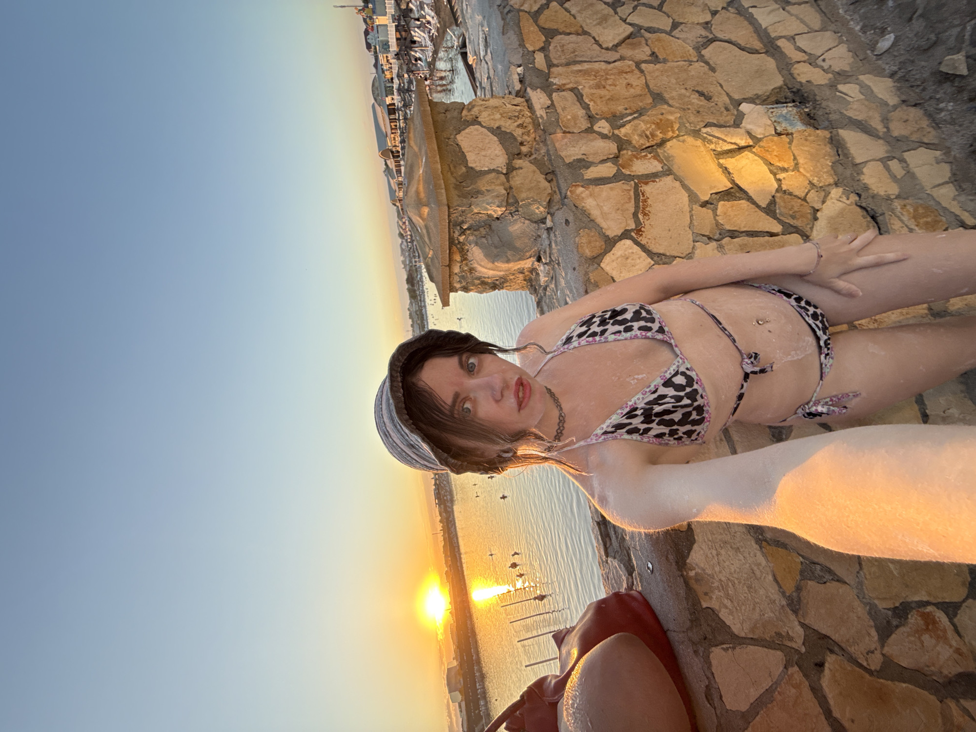 The image captures a young woman posing for a selfie at sunset by a body of water, likely in Sol-Iletsk, Russia, known for its salt lakes. She is wearing a leopard-print bikini and a bucket hat. The setting appears to be a resort or beach area, with buildings and beach chairs visible in the background. The sun is low on the horizon, casting a warm glow over the scene. The woman's skin is dusted with a white substance, possibly salt from the lake, which is a common practice in Sol-Iletsk for therapeutic reasons. The foreground features a stone wall and a person's leg to the left of the frame.