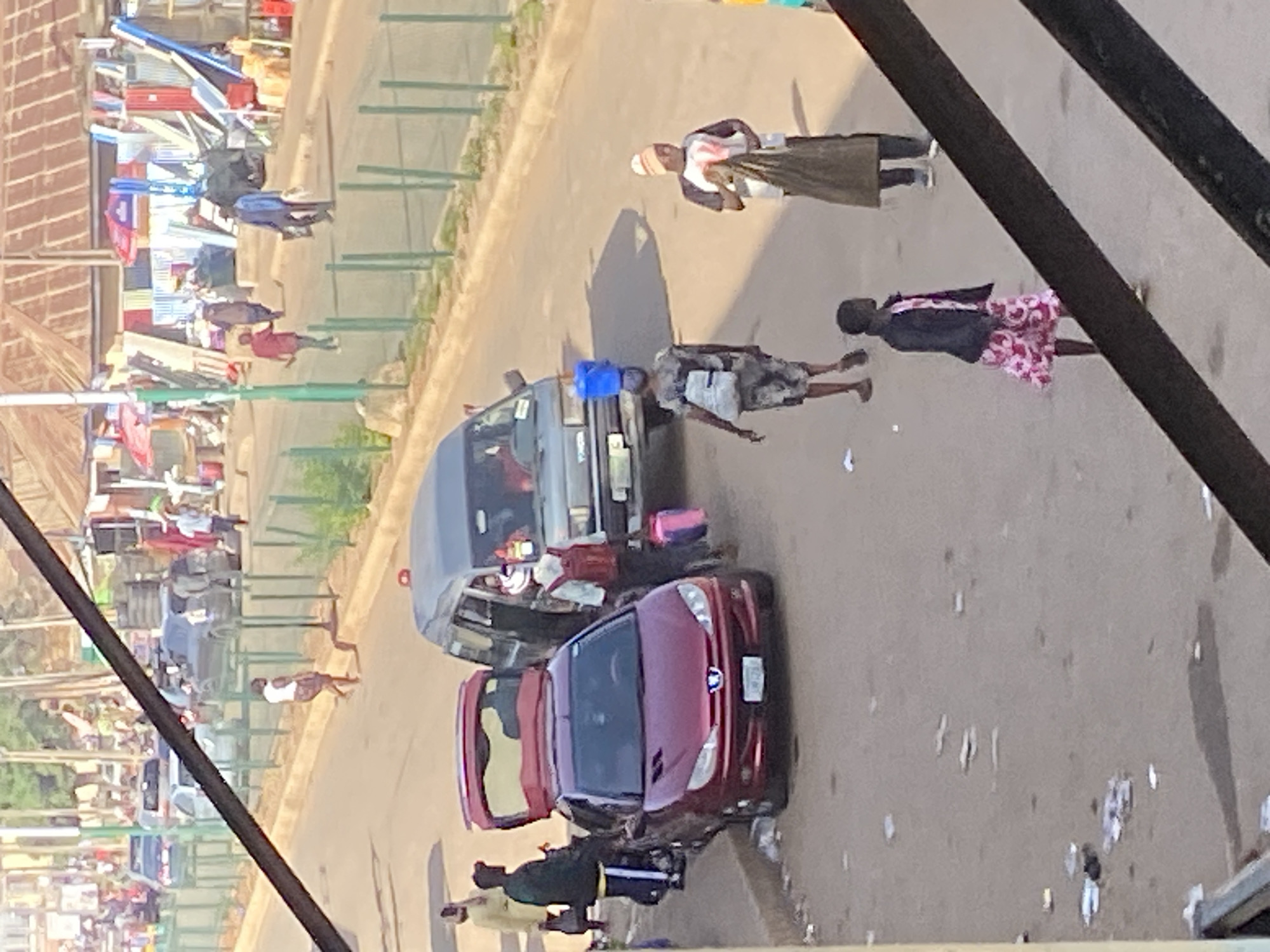 In Bukuru, Nigeria, a street scene unfolds with several individuals and vehicles. A burgundy Peugeot 206 sedan is parked at the curb with its trunk open, and a maroon minivan is also present. Two men, one in a dark jacket and the other in a lighter shirt and dark pants, stand near the Peugeot. A woman in a white top and patterned skirt walks past the vehicles. Further down the street, a man in a blue jacket carries a bag. To the right, a person wearing a patterned outfit walks away from the viewer, and another individual in a white and black shirt and a beanie is holding a large brown bag. A woman with a backpack carries a blue water container on her head, walking near the minivan. The environment appears to be a bustling street with buildings and shops in the background, and a green fence lines the edge of the road. Scattered litter is visible on the ground.