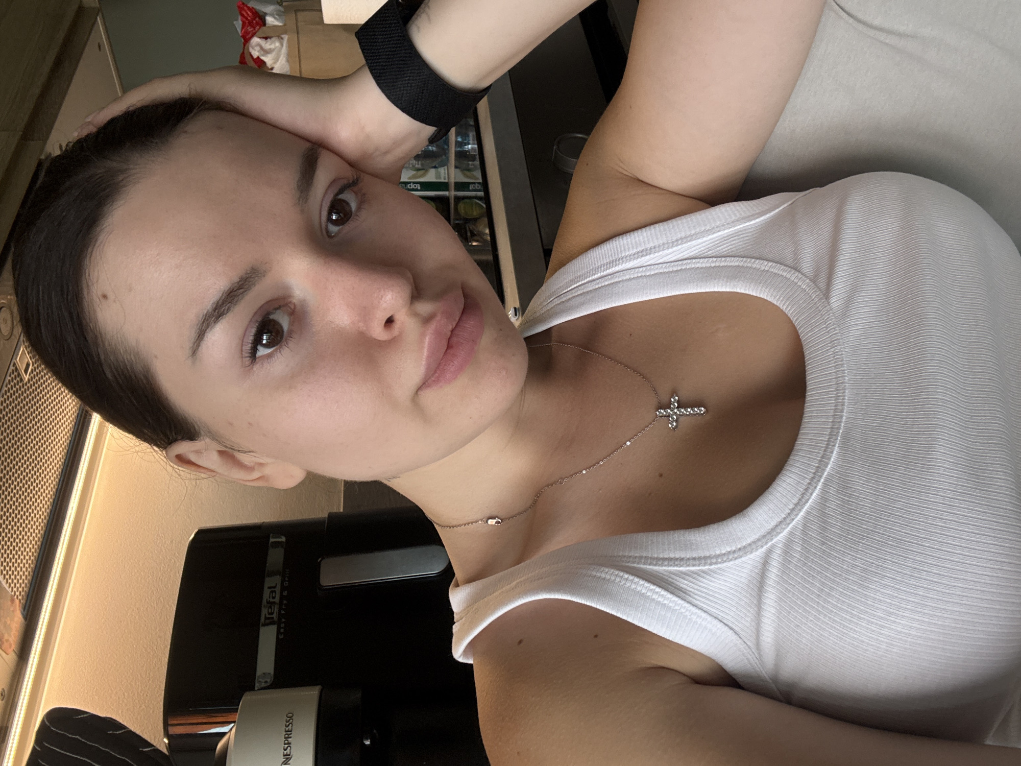 The image shows a close-up, selfie-style shot of a young adult female resting her head on her right hand, with her left arm resting on her lap. She has dark brown hair pulled back, brown eyes, and fair skin. She is wearing a white ribbed tank top and a silver necklace with a jeweled cross pendant. A black watch or fitness tracker is visible on her right wrist.
In the background, a black kitchen appliance, possibly an air fryer, with a "Tefal" logo is visible, along with a Nespresso coffee machine. The environment appears to be a kitchen or dining area, with kitchen cabinets and a countertop visible. The lighting is warm and indoor. The image does not provide any specific visual cues to indicate the location is in Prague, Czechia.