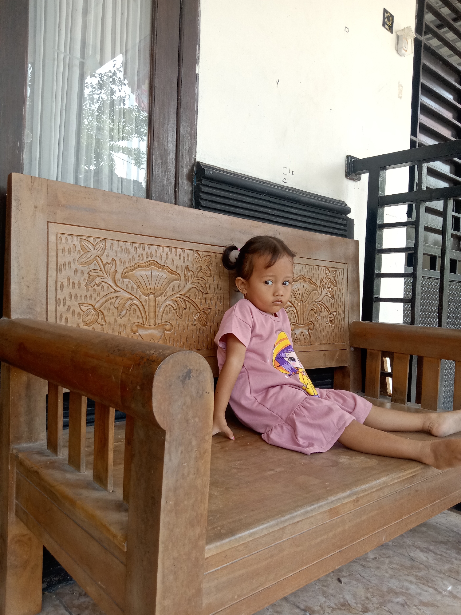 A young girl with dark hair in two small ponytails, wearing a pink dress with a graphic of a cartoon character on the front, sits on a wooden bench. The bench is made of light-colored wood and features intricate carved floral designs on the backrest. The girl is looking to her left, away from the camera, with a slightly pouty expression. Her legs are extended in front of her, and her feet are bare. The background shows a white wall, a window with white curtains, and a black metal fence. The scene appears to be outdoors, possibly on a porch or veranda, with a tiled floor visible beneath the bench. This appears to be in a residential area in Stiaji, Indonesia.