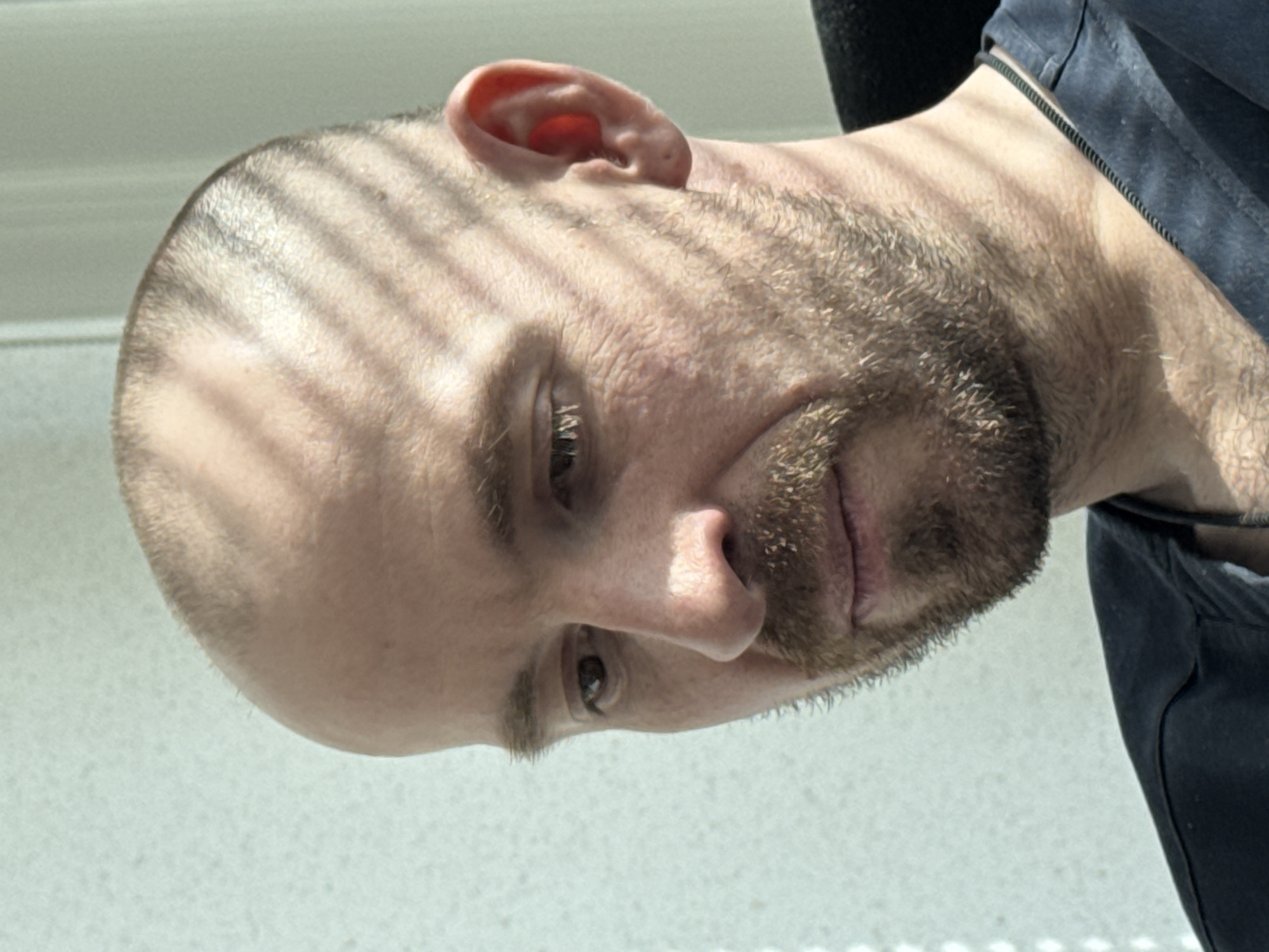A close-up, tilted headshot of a bald, bearded man is shown. The man is facing toward the bottom left of the frame. A shadow cast by window blinds falls across the top of his head. The man is wearing a dark-colored shirt. The background appears to be a solid, light-colored surface.
The location cannot be verified as Прикамский, Russia, based solely on the visual information provided.
