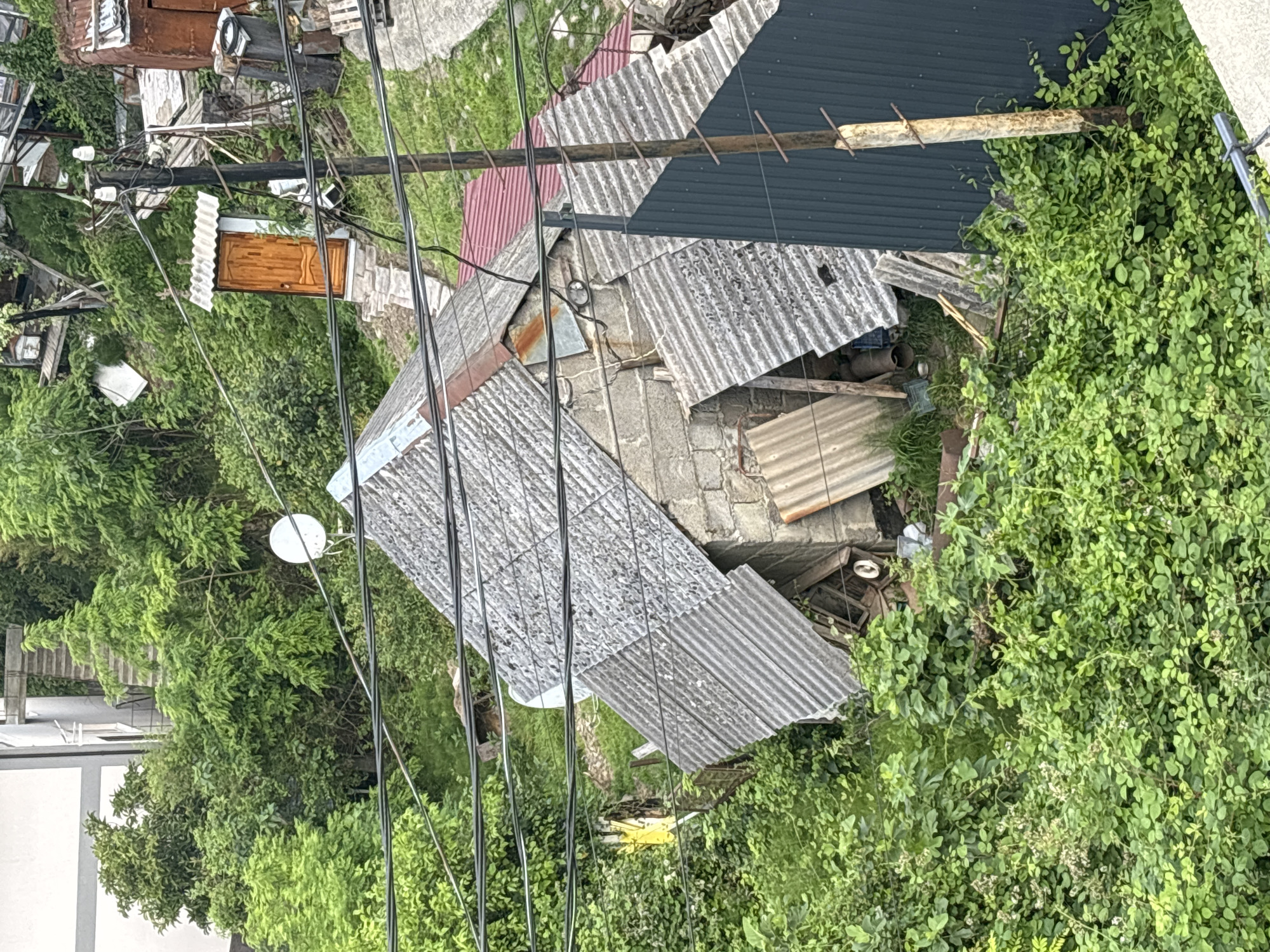 A high-angle, full shot shows a dilapidated structure with a corrugated metal roof and cinder block walls, overgrown with lush green vegetation. The building is situated on a sloped, uneven terrain, possibly a hillside. Numerous black power lines crisscross the upper portion of the frame, strung between wooden utility poles. To the right of the main structure, a small wooden outhouse with a wooden door and a corrugated roof sits on a stone foundation. In the background, a large white satellite dish is visible amongst the dense foliage. Various pieces of debris, including corrugated sheets of metal and pipes, are scattered around the property. The overall impression is one of disrepair and neglect, with nature encroaching upon the man-made structures. The environment appears to be a residential area, though the specific location is not identifiable.