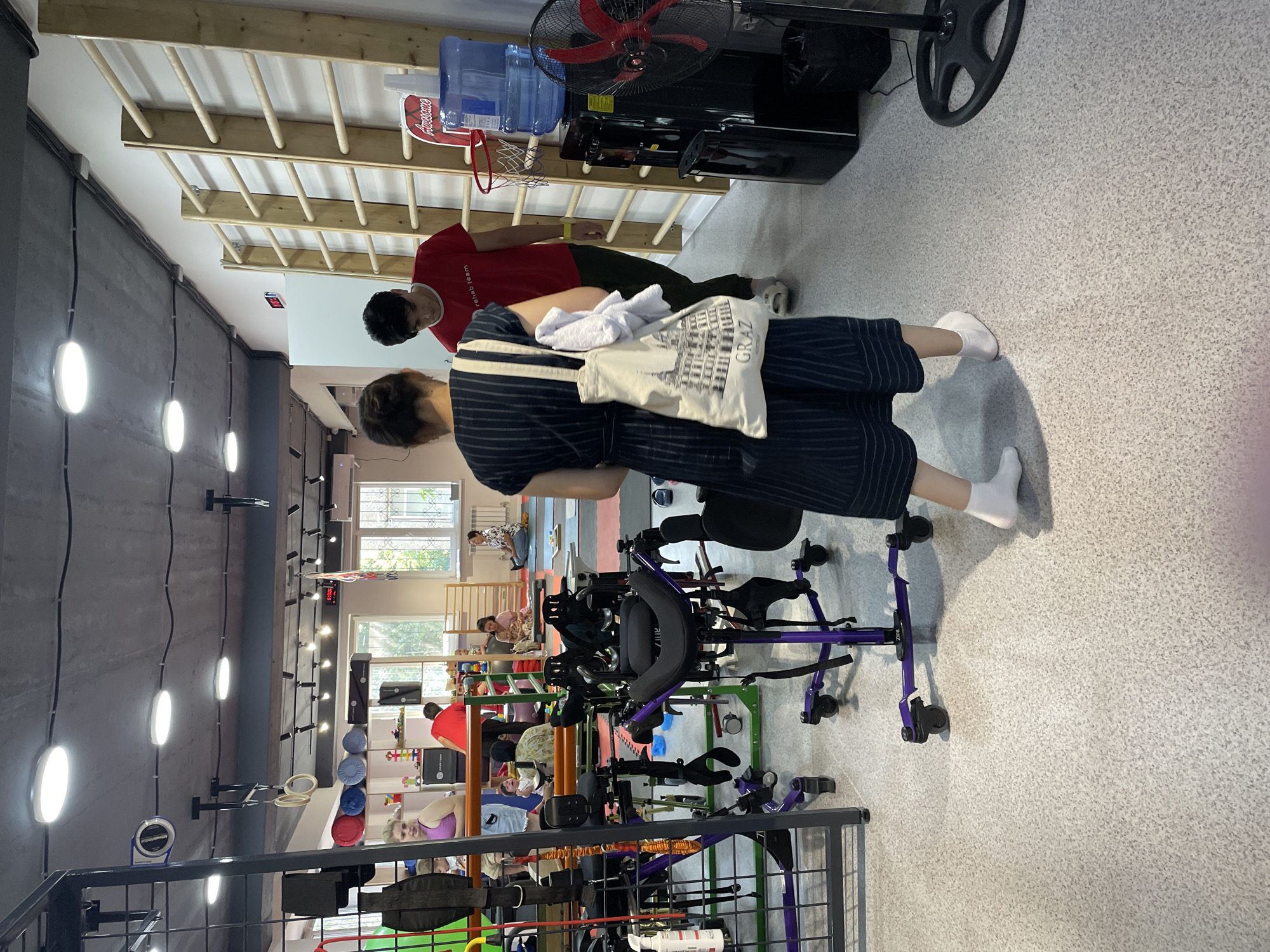 The image depicts the interior of a rehabilitation or therapy center, likely in Almaty, Kazakhstan, given the context. The room is well-lit with overhead fluorescent lights and features a textured, light-colored speckled floor. The space is equipped with various therapeutic and exercise equipment, including a purple walker or gait trainer with a seat, a large metal frame with straps, gymnastic rings hanging from the ceiling, and a wooden wall ladder with horizontal bars. In the background, there are windows, a water cooler with a large bottle of water, a red basketball hoop, and a black oscillating fan.
Several people are present in the scene. In the foreground, a woman wearing a dark blue pinstripe dress and white socks is walking away from the camera, carrying a tote bag with an image of buildings and the word "GRAZ" on it. To her right, a young man wearing a red t-shirt that reads "rehab team" and dark pants is standing near the wall ladder. In the middle ground, a woman in a purple tank top and denim shorts is looking towards the left, with children visible behind her. Another woman is seated on the floor in the background, with a child in her lap. The overall atmosphere suggests a functional and active environment designed for rehabilitation and exercise.