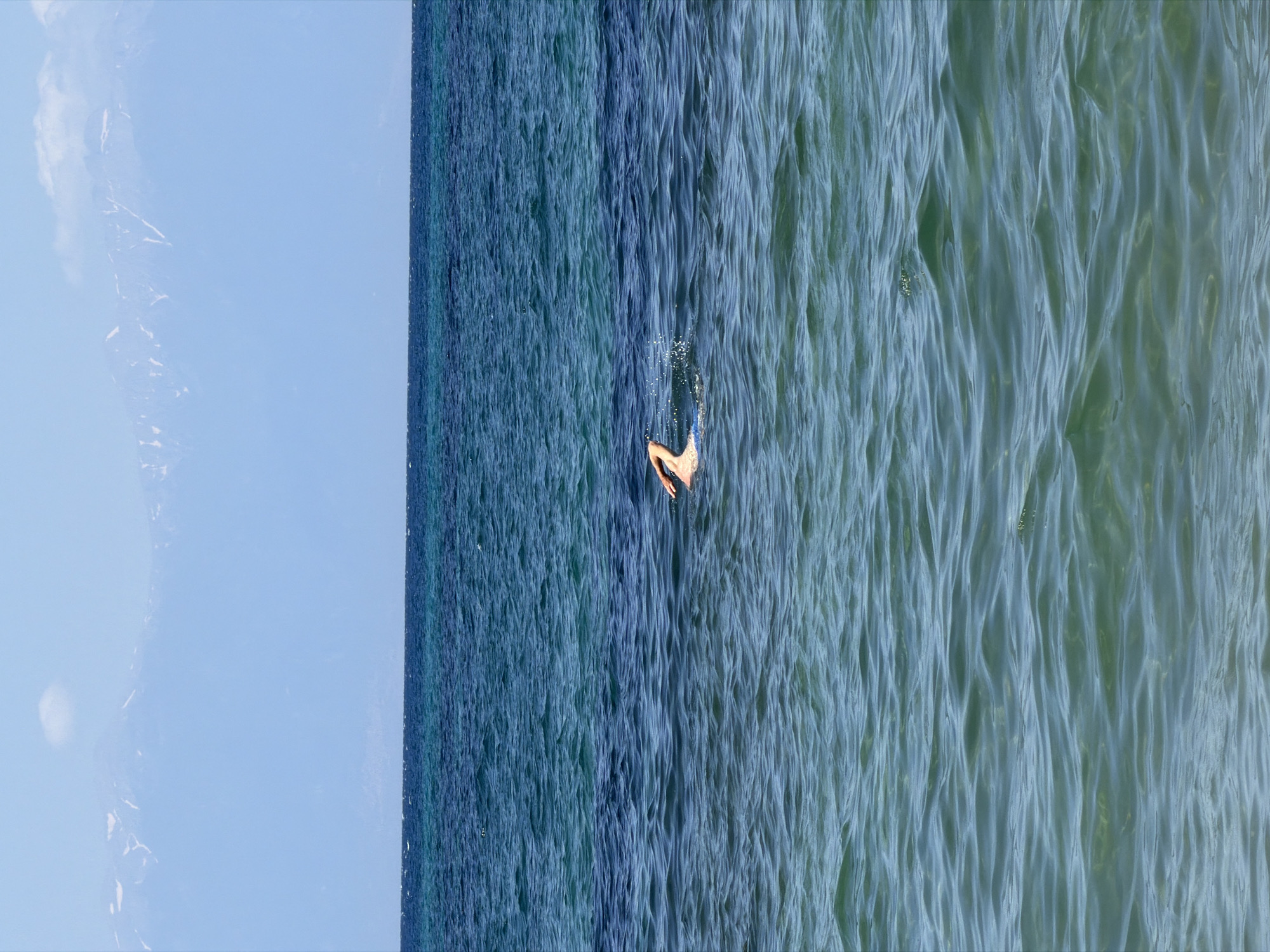 A person is swimming in the sea. The water is a deep blue, transitioning to a lighter, more translucent turquoise closer to the foreground. The surface of the water is rippled, indicating a gentle current. In the background, a distant mountain range is visible, its peaks dusted with snow under a clear blue sky. The scene appears to be located near Νέα Ηράκλεια, Greece, given the distinct view of Mount Olympus across the Thermaic Gulf.