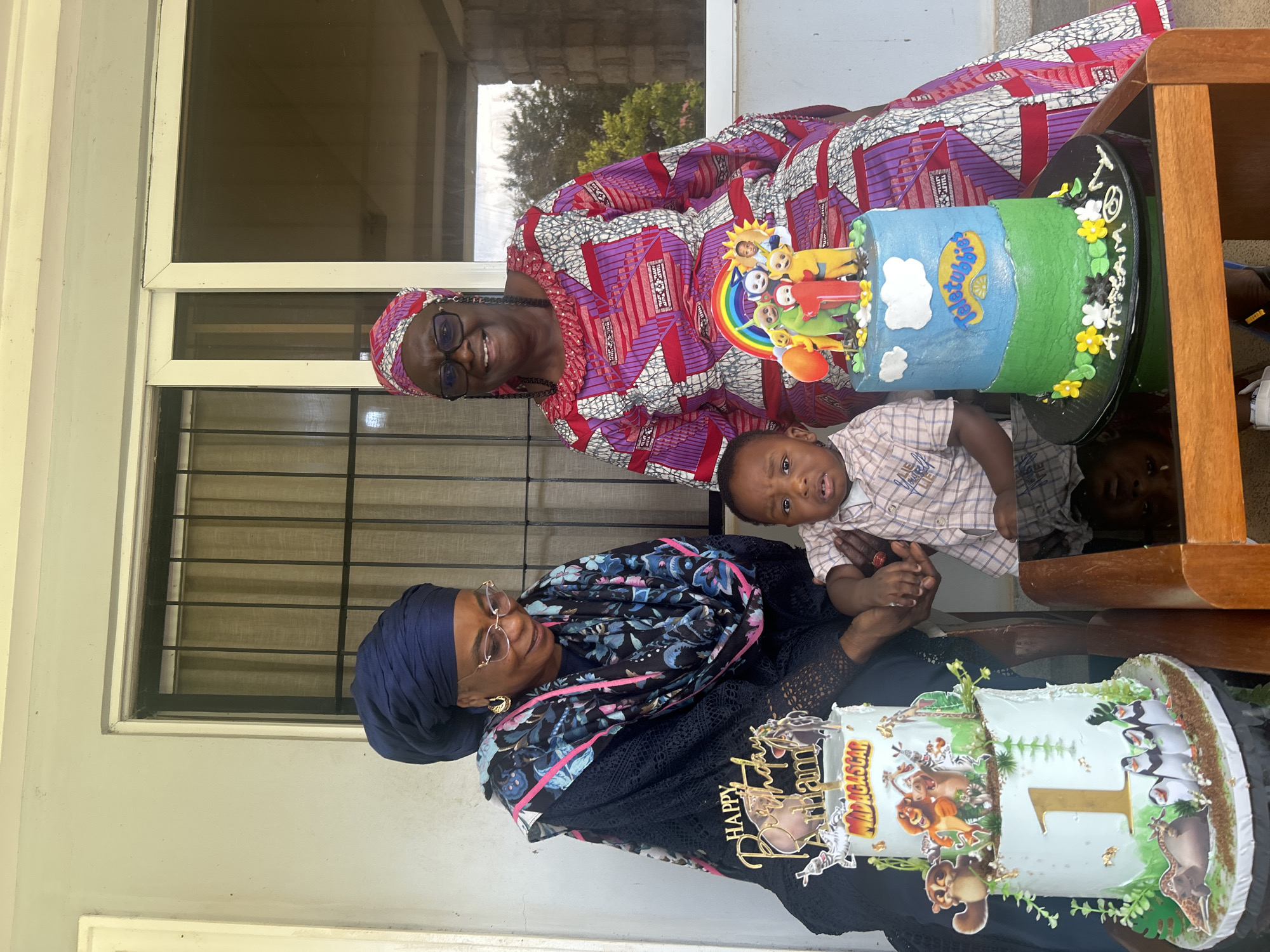 The image shows a frontal, medium shot of two women and a young child sitting on a porch. The woman on the left wears a dark blue headscarf and a black patterned dress with a lacy overlay. She is wearing glasses and golden earrings. The woman on the right wears a pink and red patterned headscarf and a matching dress. She is also wearing glasses and earrings. Between them sits a young child wearing a plaid shirt. In front of the women are two cakes. The cake on the left is a two-tiered cake decorated with characters from Madagascar and a large "1" on the bottom tier. The cake on the right is decorated with characters from Teletubbies and a rainbow. The porch appears to be made of concrete, with a railing visible behind the subjects. The background shows a window with vertical blinds and some foliage visible outside. The scene is lit by natural light. The location is the city of Dutsen Kato, Nigeria.