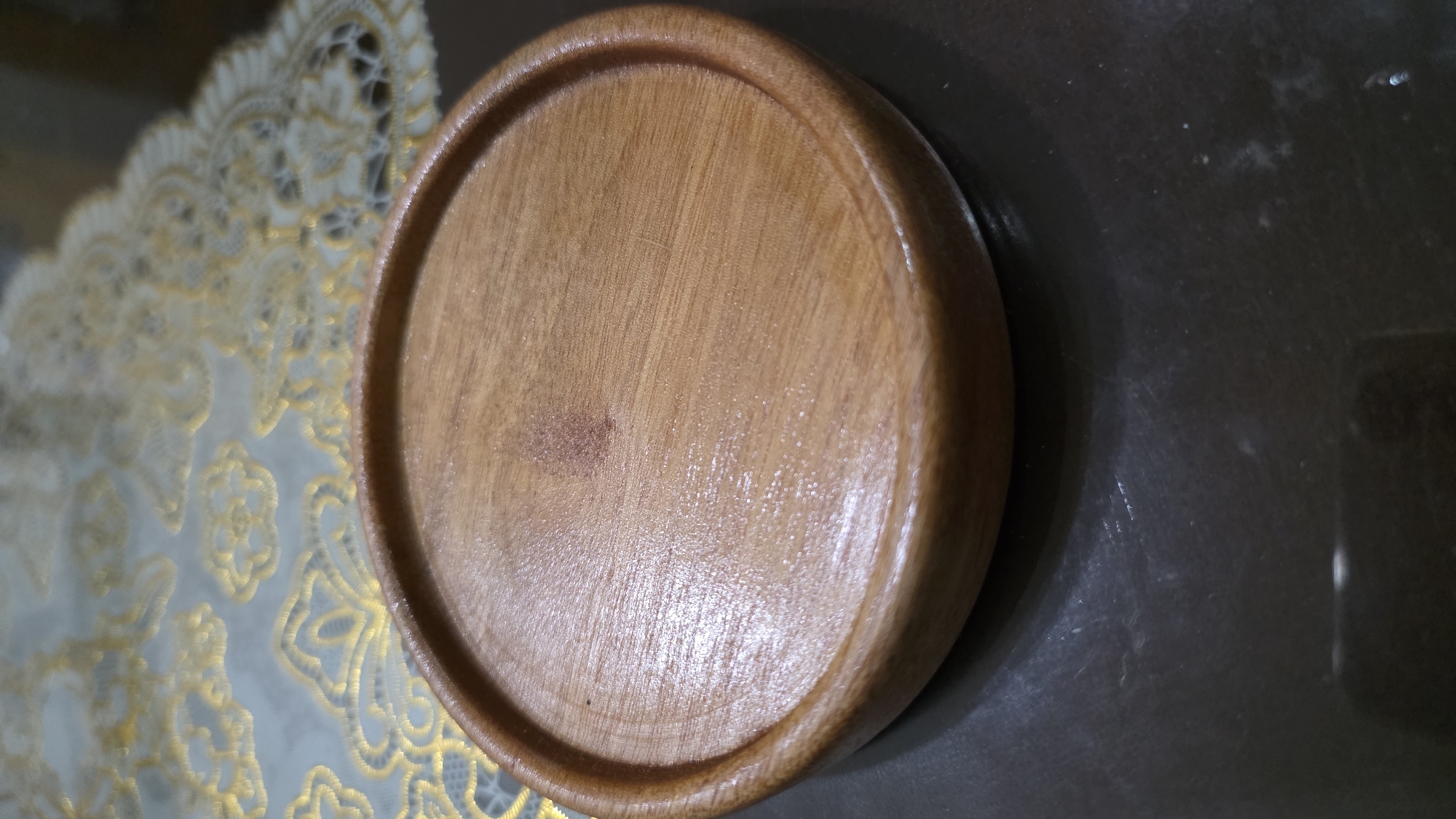 A circular wooden coaster sits on a dark, reflective surface. The coaster has a raised outer edge and a slightly concave inner surface. The wood grain is visible, and there is a small, darker stain or mark on the upper portion of the coaster. Behind the coaster, a light-colored tablecloth with an ornate gold pattern is visible. The scene is lit from above, creating highlights on the coaster's surface. There are no people or any other objects clearly identifiable in the image. The location is identified as Pondok Kaso Tonggoh, Indonesia.