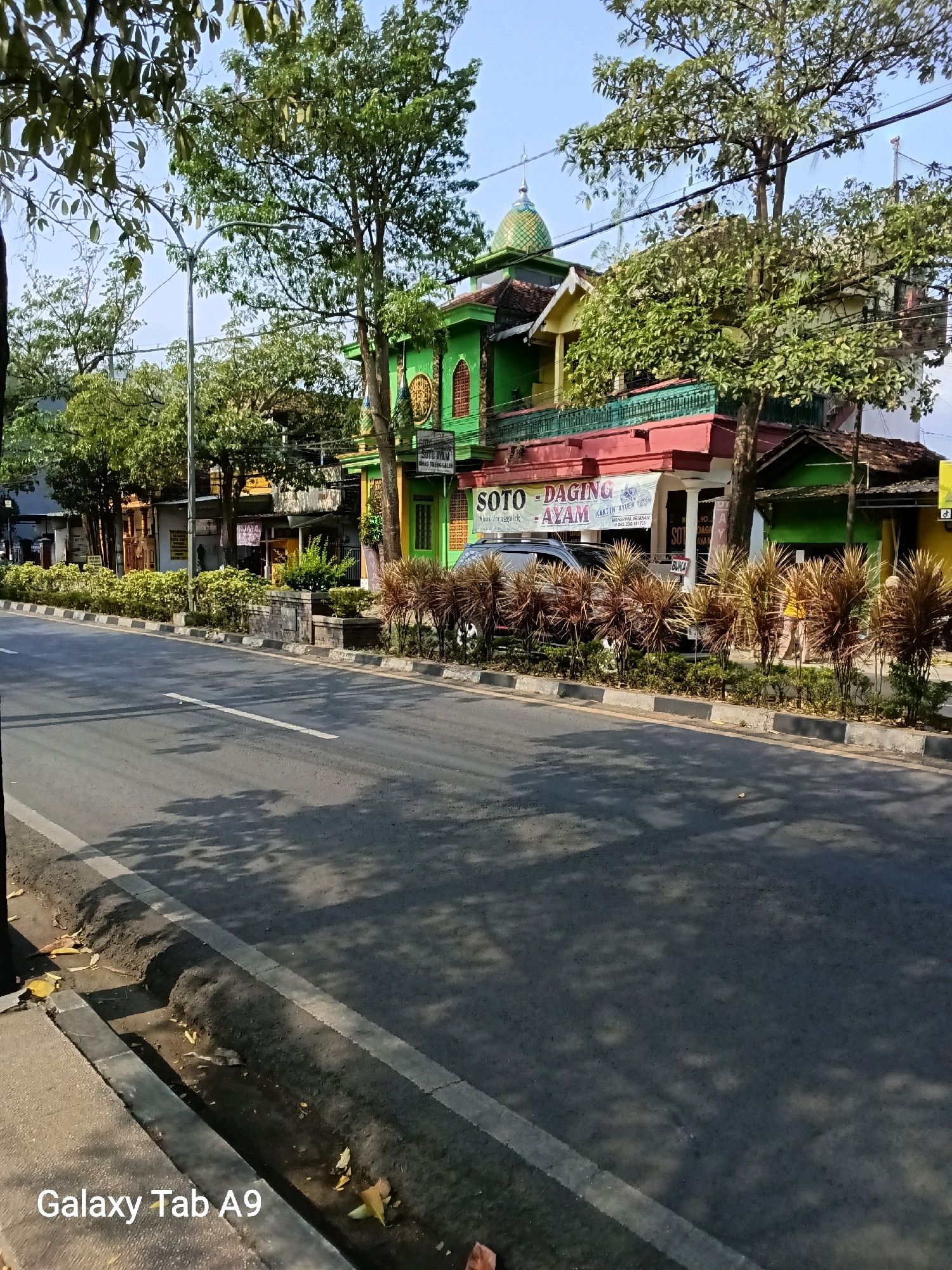 A street scene in Kelutan, Indonesia, depicts a road with a sidewalk and a row of trees lining the buildings. The buildings are colorful, with a prominent green and red structure housing a food establishment that advertises "SOTO - DAGING AYAM" (Chicken Meat Soup). A dark-colored SUV is parked in front of the establishment. The road is paved and marked with a white line. Shadows from trees are cast across the road. The environment appears to be sunny. The image is captured from a low angle, showing the bottom of a dark pole on the left side and a sidewalk. The text "Galaxy Tab A9" is present in the bottom left corner.