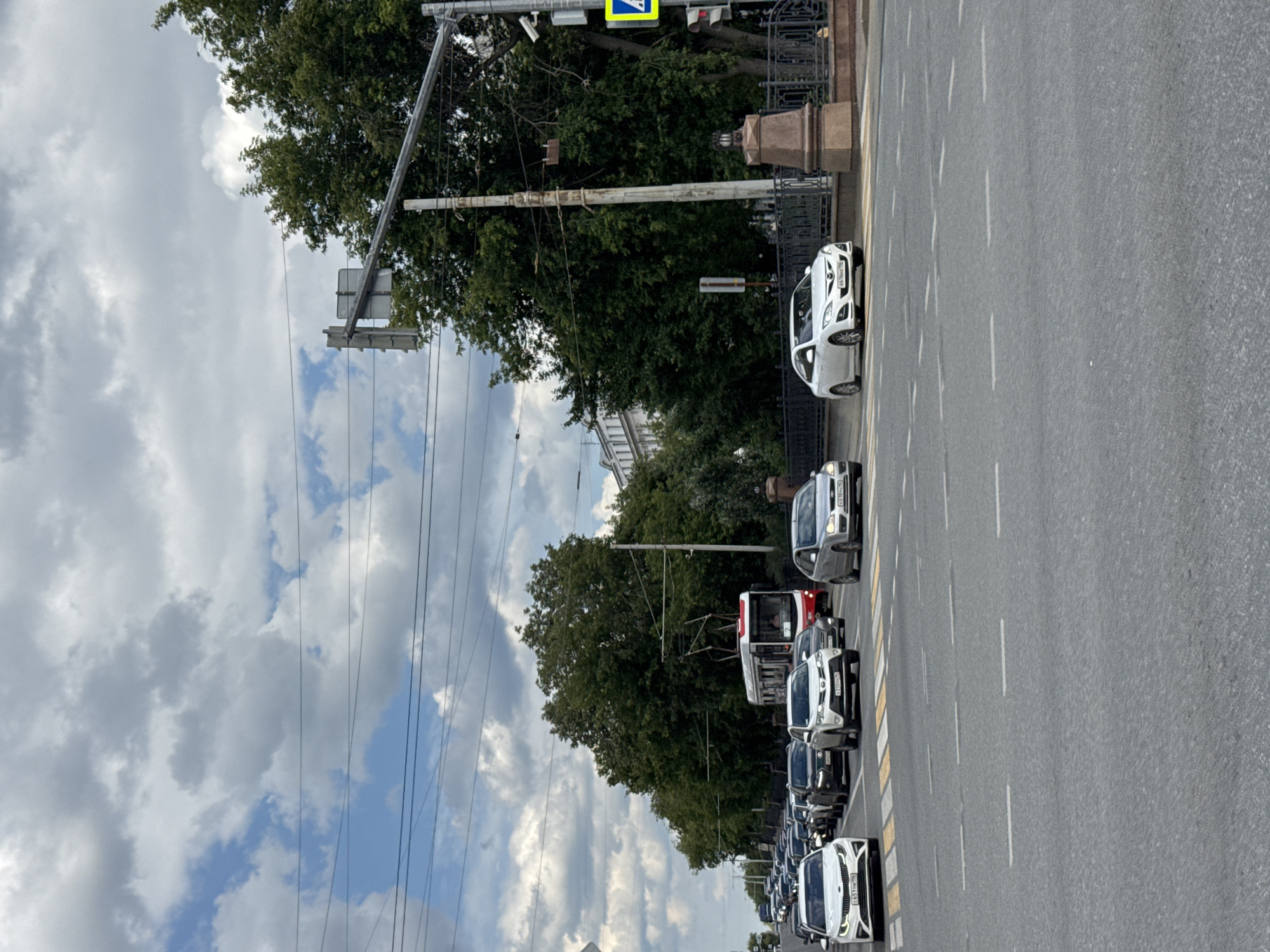 In Yekaterinburg, Russia, on a cloudy day, traffic moves along a multi-lane asphalt road. Cars of various makes and models are visible, including a white Kia, a silver sedan, a white SUV, and a white sedan. Further down the road, a red and white tram is in motion, surrounded by a queue of cars. Trees line the street on the right side, and a pedestrian crossing is marked on the road. Traffic lights and streetlights are present, along with a security camera mounted on a pole. The sky is filled with a mix of gray and white clouds, with patches of blue sky visible. Buildings are visible in the distance, suggesting an urban environment.