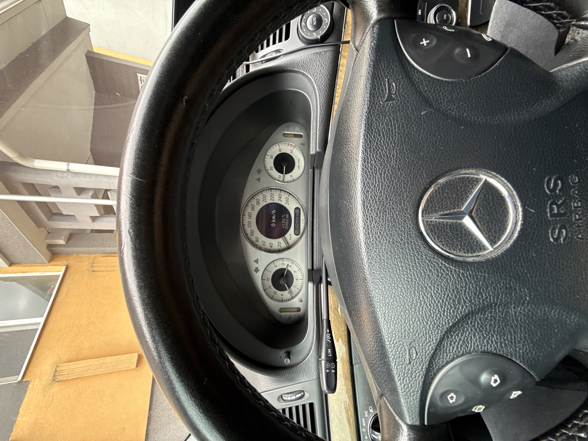 The image shows the dashboard and steering wheel of a Mercedes-Benz car. The speedometer reads 0 km/h, and the odometer displays 896.0 km. The steering wheel has the Mercedes-Benz logo and various controls. The outside temperature is 13.0°C. Through the windshield, a view of a building with a window and a yellow wall is visible. Based on the image details, a precise location in the city of Stara Zagora, Bulgaria, cannot be determined.