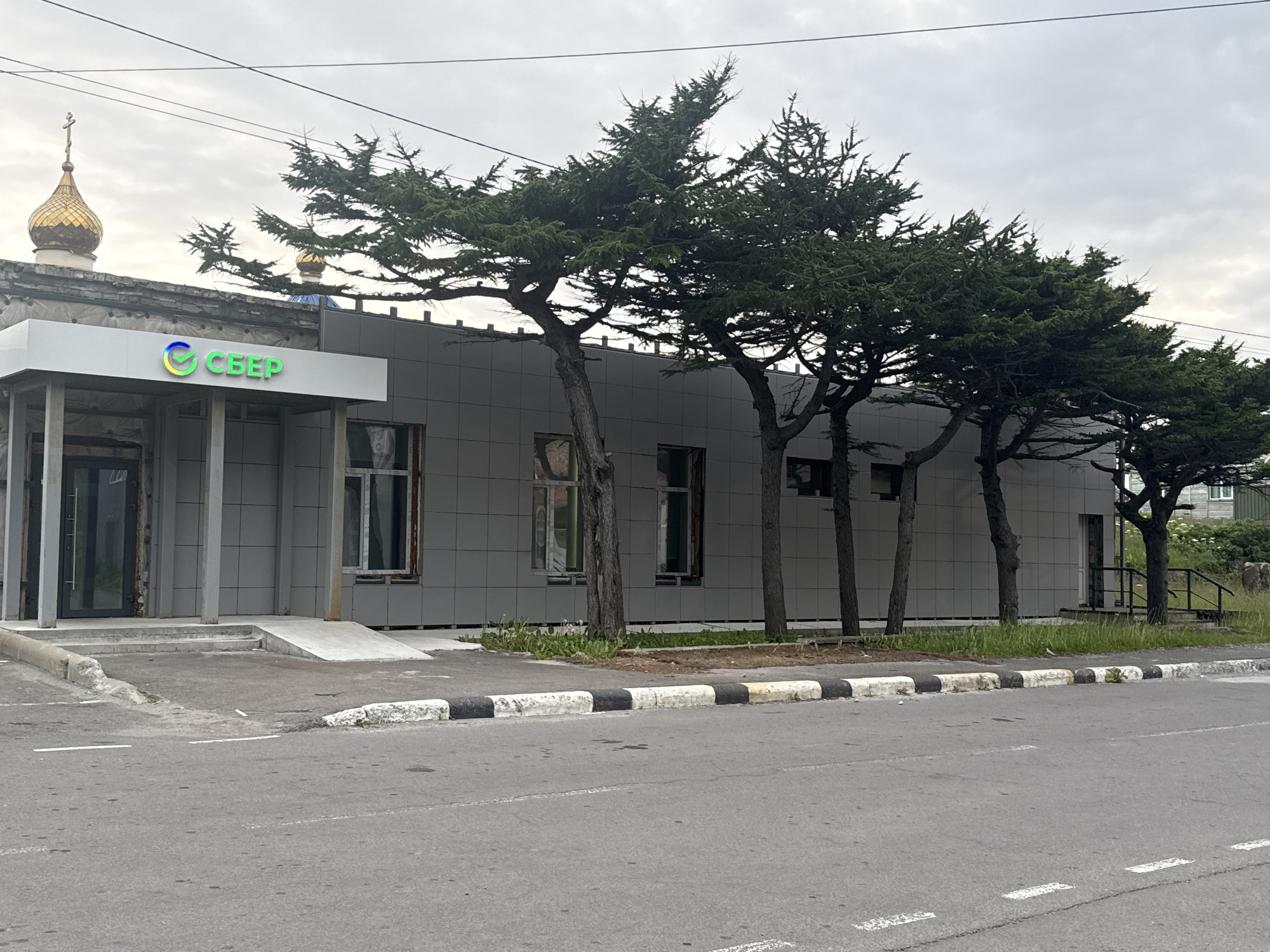 The image shows a modern building with a gray tiled facade, featuring a prominent green and yellow logo that reads "СБЕР" (Sber). The building has large windows, some of which appear to be under construction or boarded up. In front of the building, there is a sidewalk with a ramp for accessibility. Several coniferous trees with distinct, somewhat windswept branches are planted along the front of the building, creating a natural barrier.
To the left of the Sber building, a golden-domed Orthodox church steeple is visible, suggesting a mix of modern and historical architecture in the vicinity. The street in front of the building is asphalt, marked with white lines, and bordered by a curb painted with alternating black and white stripes. The sky is overcast, indicating cloudy weather. The overall environment suggests a commercial area within the city of Kuril'sk, Russia, likely a branch of Sberbank given the prominent signage. There are no people visible in the image. The scene appears to be captured during daylight hours.
