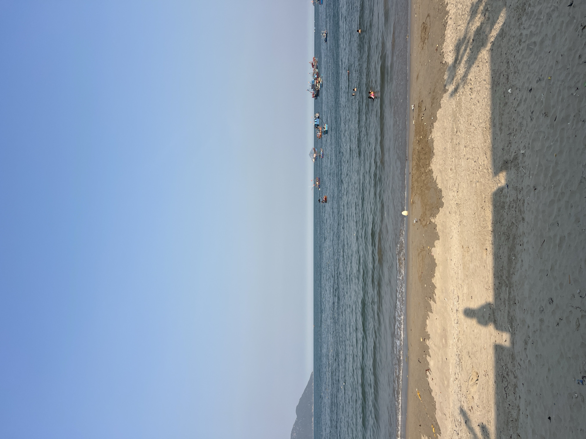 The image shows a beach scene in Vĩnh Lương, Vietnam, with a light blue sky dominating the left side. The ocean occupies the central portion of the image, with several boats and people visible in the water. The beach extends along the right side, with shadows of people cast on the sand. Debris and small objects are scattered on the beach. A small land formation is visible in the distance.