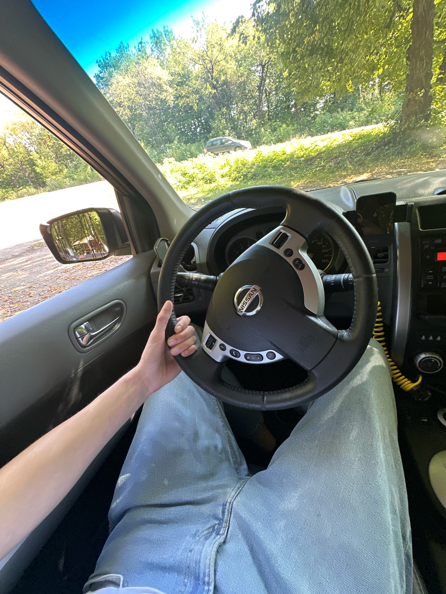 A first-person perspective from inside a car shows a person's left arm and hand resting on the steering wheel. The person is wearing light blue jeans. The car's interior is visible, including the steering wheel with a Nissan logo, dashboard, door panel, and side mirror. Outside the car, through the windshield and side window, a tree-lined area with a road and another car in the distance is visible. The scene appears to be a daytime outdoor setting.