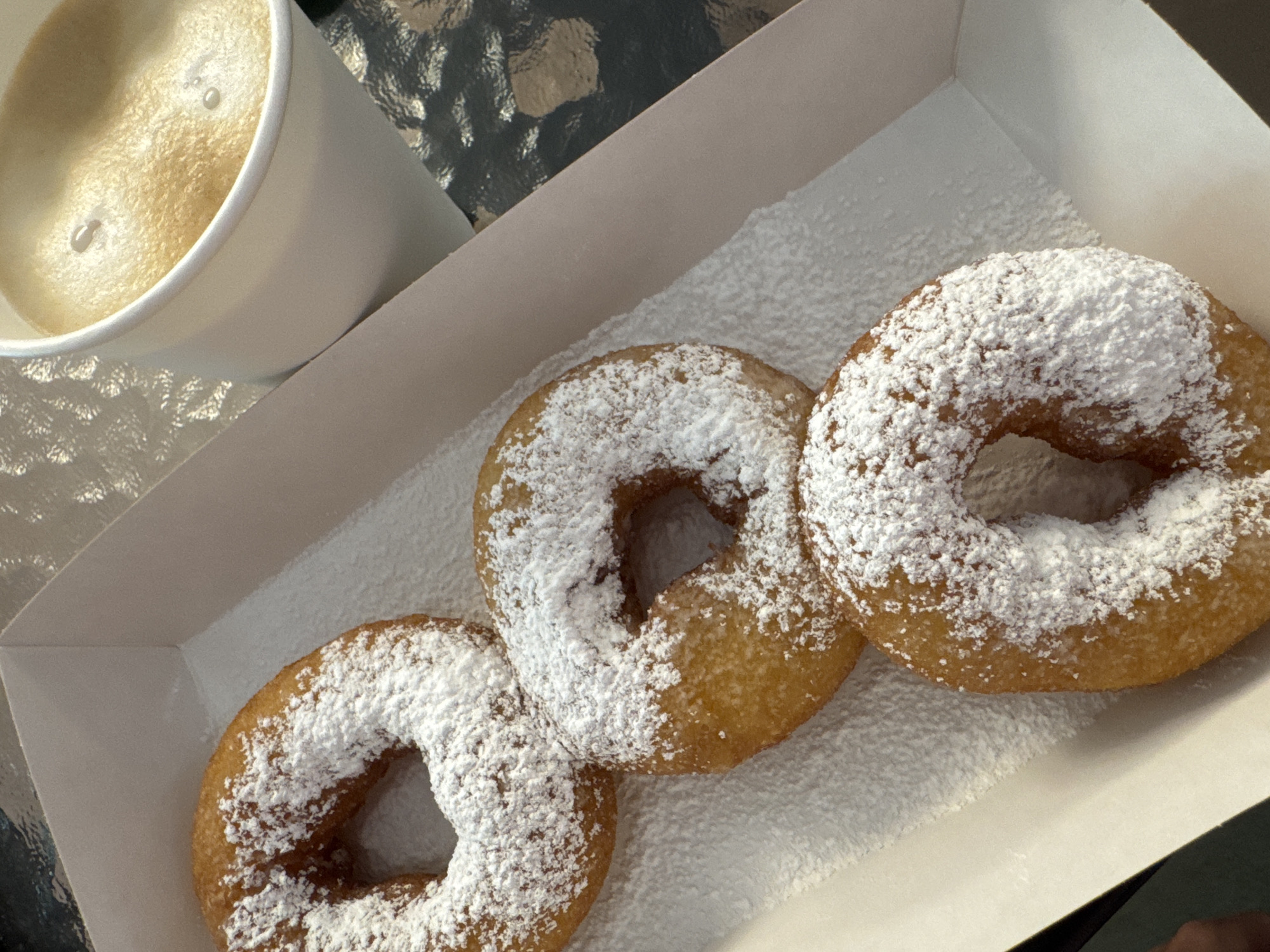 This close-up, overhead shot displays three powdered sugar donuts and a cup of coffee, likely part of a breakfast or snack. The scene is set outdoors, with the reflective, textured surface of a table visible beneath and around the items, possibly in Saint Petersburg, Russia.
The main subjects are three ring-shaped donuts, golden-brown and generously coated with white powdered sugar, creating a snowy appearance. They are arranged within a simple white paper box, which is also dusted with a fine layer of sugar. To the left and slightly behind the donuts, a white paper cup holds a light brown beverage, presumably coffee, topped with a frothy layer and a couple of visible bubbles.
The image suggests a peaceful moment, perhaps enjoying a sweet treat with a hot drink. The lighting indicates it is daytime, possibly morning or early afternoon. No people are visible, and there is no discernible text within the image. The focus is entirely on the food and drink, highlighting their appetizing textures and colors.