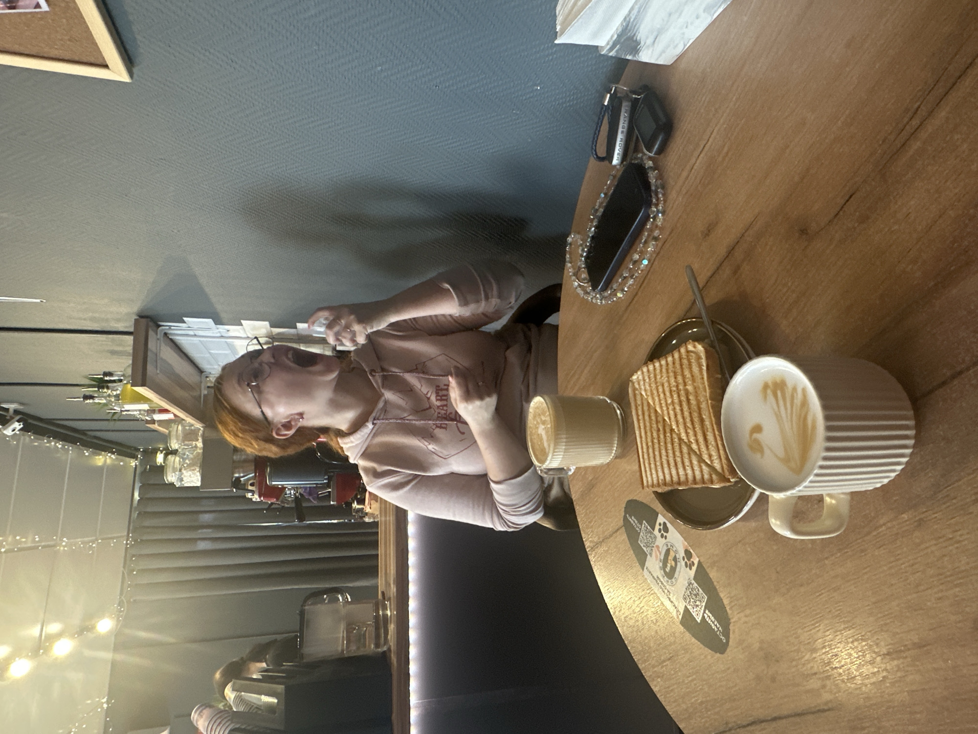 The image shows a woman sitting at a wooden table in what appears to be a cafe or restaurant setting. The table is set with a plate of toast, two cups of coffee (one with latte art), and a business card. The woman is fair-skinned with red hair, wearing glasses and a light pink hoodie. She is holding a phone or small device in her hand. On the table next to her are a cell phone and a set of car keys. The background shows a bar area with liquor bottles and other cafe equipment. The location seems to be Unknown, Unknown.