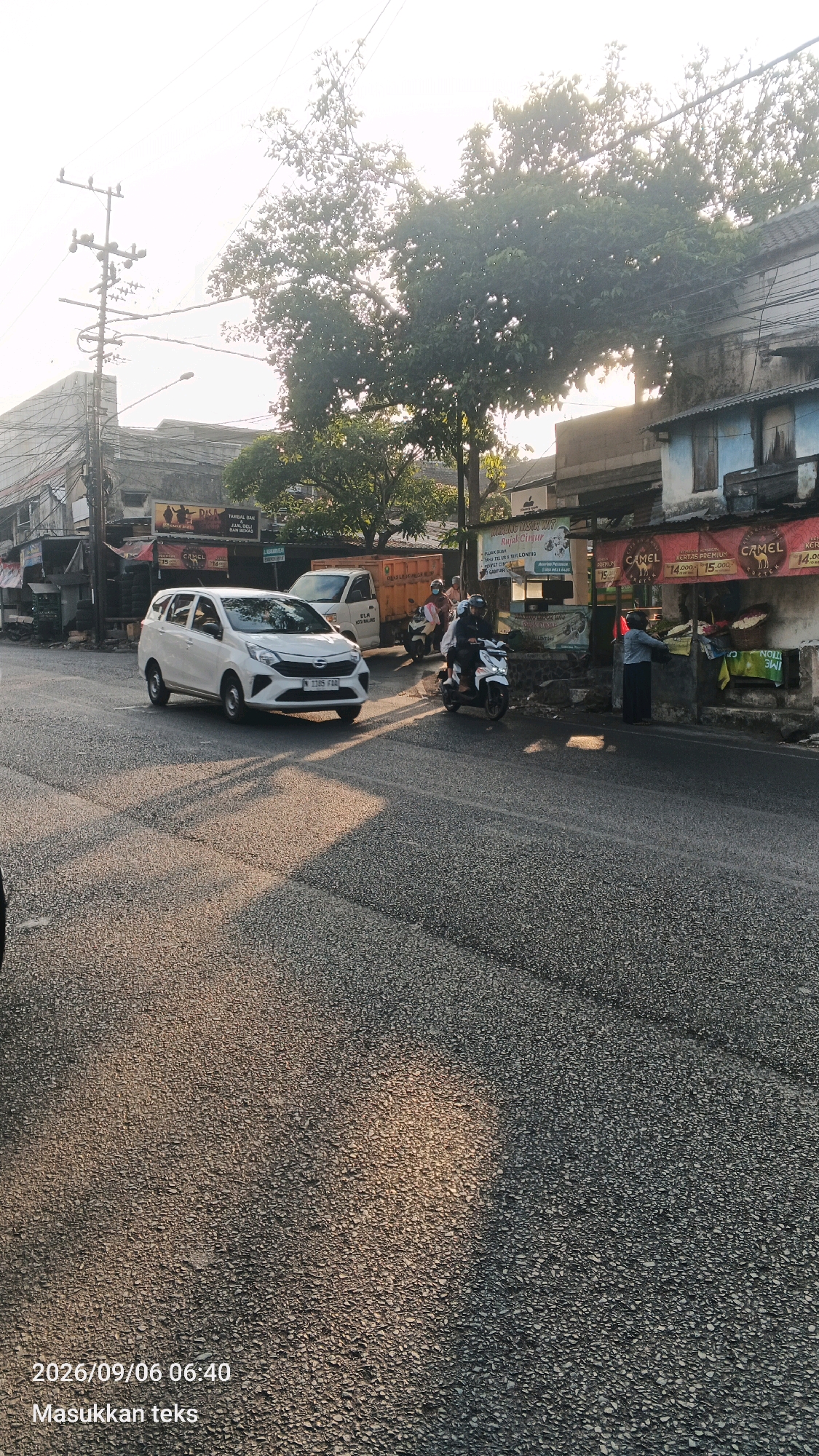 In Malang, Indonesia, a white car drives down a street lined with buildings and trees. The sun casts long shadows across the asphalt road. On the right side of the street, a food stall is set up under a canopy, with a woman attending to customers. A man on a motorcycle passes by the stall, and another motorcycle is parked nearby. A truck is visible further down the street, and a utility pole with multiple wires stands on the left side of the road. The sky is bright and overcast, suggesting it is either early morning or late afternoon. The overall scene depicts a typical day on a street in a bustling Indonesian city.
