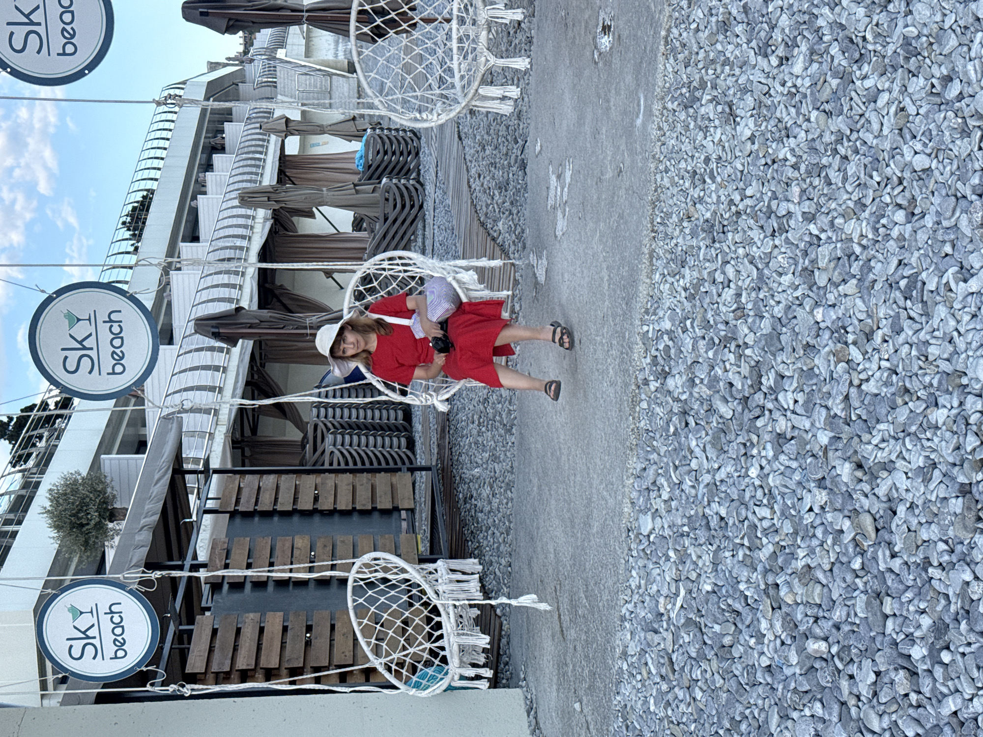 A young woman in a red dress and a white hat sits in a white macrame hanging chair. She is holding a camera and a small purse is slung over her shoulder. She is wearing dark sandals. The setting is outdoors on a paved walkway next to a gravel area. In the background, there are tiered levels of seating with umbrellas and some hanging chairs. Two circular signs with the text "SKY beach" are visible, one hanging from a string on the left and another higher up. The sky is visible at the top of the image and appears to be daytime. The woman has a neutral expression. The location is identified as Yalta, Ukraine.