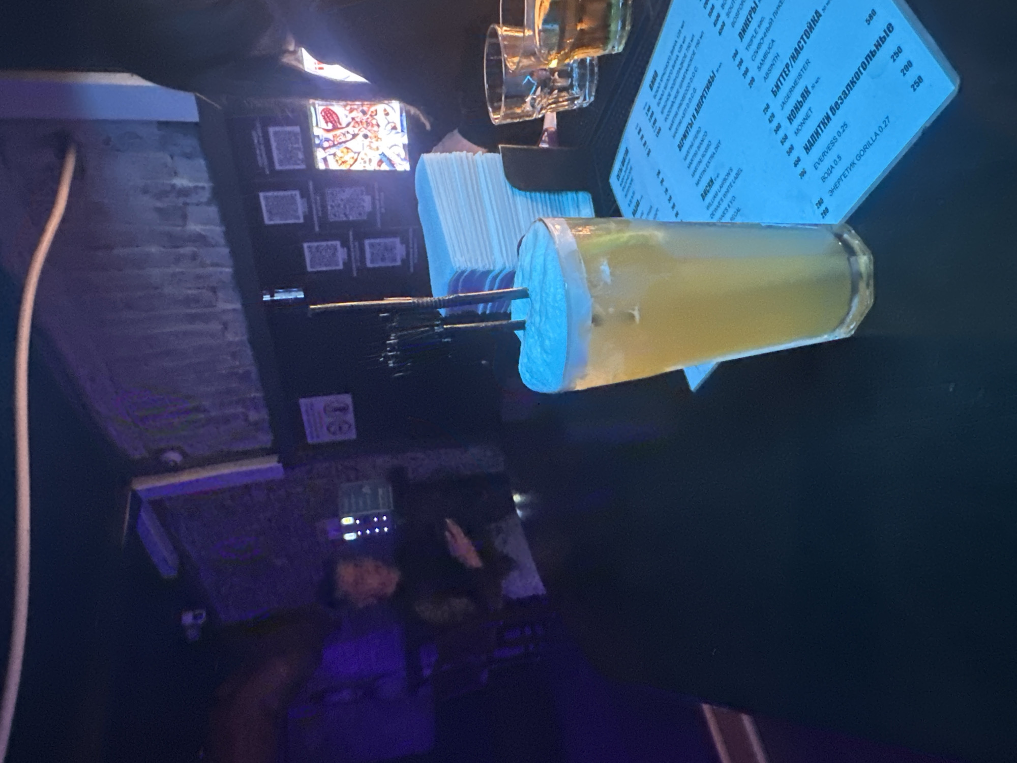A dimly lit bar or nightclub interior with purple and blue ambient lighting. In the foreground, a tall glass filled with a yellow beverage and topped with foam and two black straws sits on a dark table. Next to the drink is a stack of white napkins. To the right of the napkins are two empty glasses, one clear and one with amber liquid. A blue menu is partially visible on the table in front of the drink. In the background, a person in a dark shirt is visible, holding a phone. Behind them, a brick wall with QR codes and illuminated graphics is visible. The overall impression is of a casual night out at a bar in Perm, Russia, possibly a tourist establishment given the QR codes which might lead to menus or information in Russian.