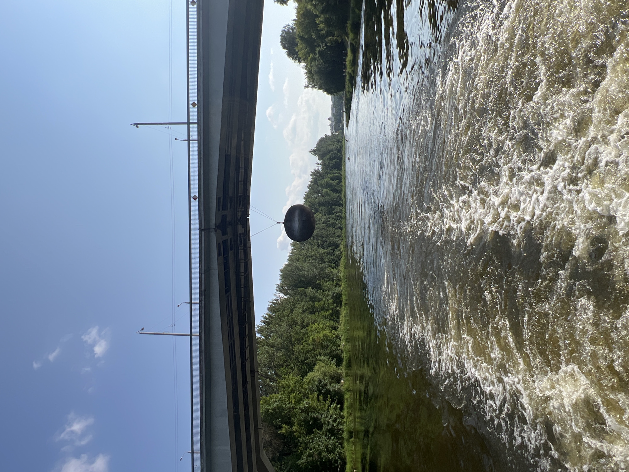 The image depicts a view from a boat traveling down a river, looking towards a large bridge. A dark, spherical object is suspended from the bridge by a thin cable. The riverbanks are lined with dense green trees. The water behind the boat shows a significant wake. The sky is blue with scattered white clouds. The bridge appears to be part of a road or railway crossing. The distant cityscape in the background suggests an urban or semi-urban environment. Given the context of the spherical art installation under the bridge, this scene is likely located in Vilnius, Lithuania, specifically on the Neris River, where the "Ball" installation by artist Gintaras Karosas is situated under the White Bridge.