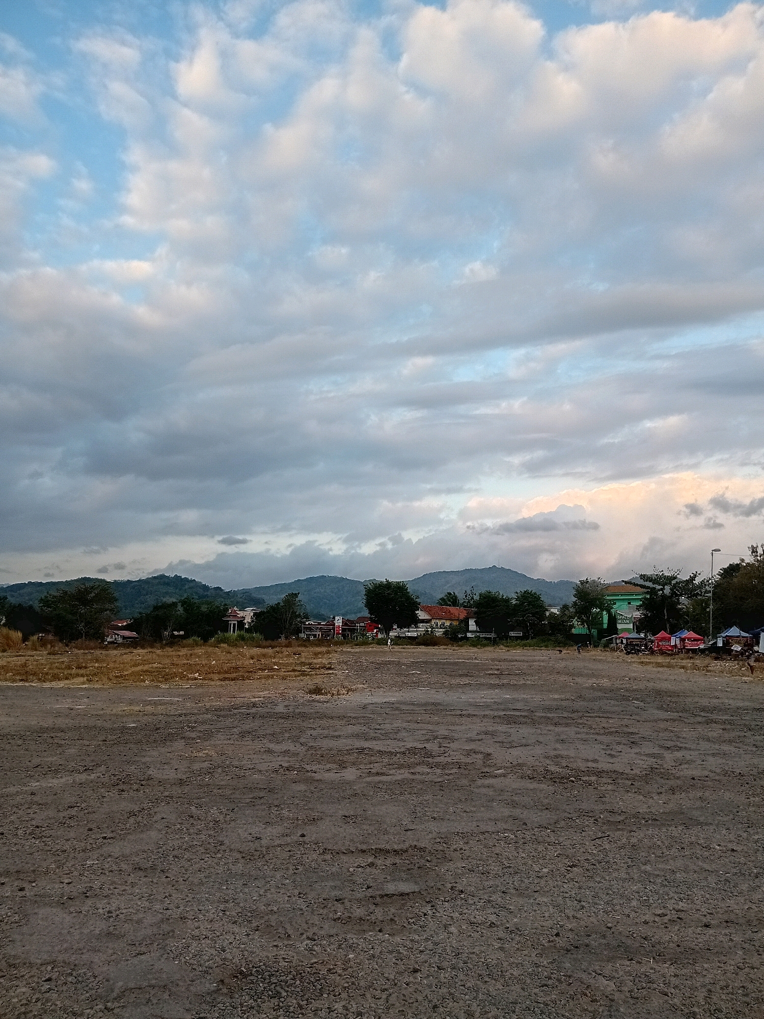 The image depicts a wide, open, dirt lot in the city of Purworejo, Indonesia. The lot is predominantly grey and textured with small stones and patches of dry, yellowed grass. In the middle ground, a line of trees separates the lot from a cluster of buildings and structures. Among these are houses with red and orange roofs, a prominent green building, and several tents or market stalls, some displaying red and blue canopies. Beyond the buildings, a range of green, tree-covered hills forms the backdrop. The sky above is a mix of light blue and dramatic, grey and white clouds, with hints of warm, soft light on the horizon, suggesting either sunrise or sunset. Several figures, appearing as small silhouettes, are visible in the distance within the lot and near the market stalls.