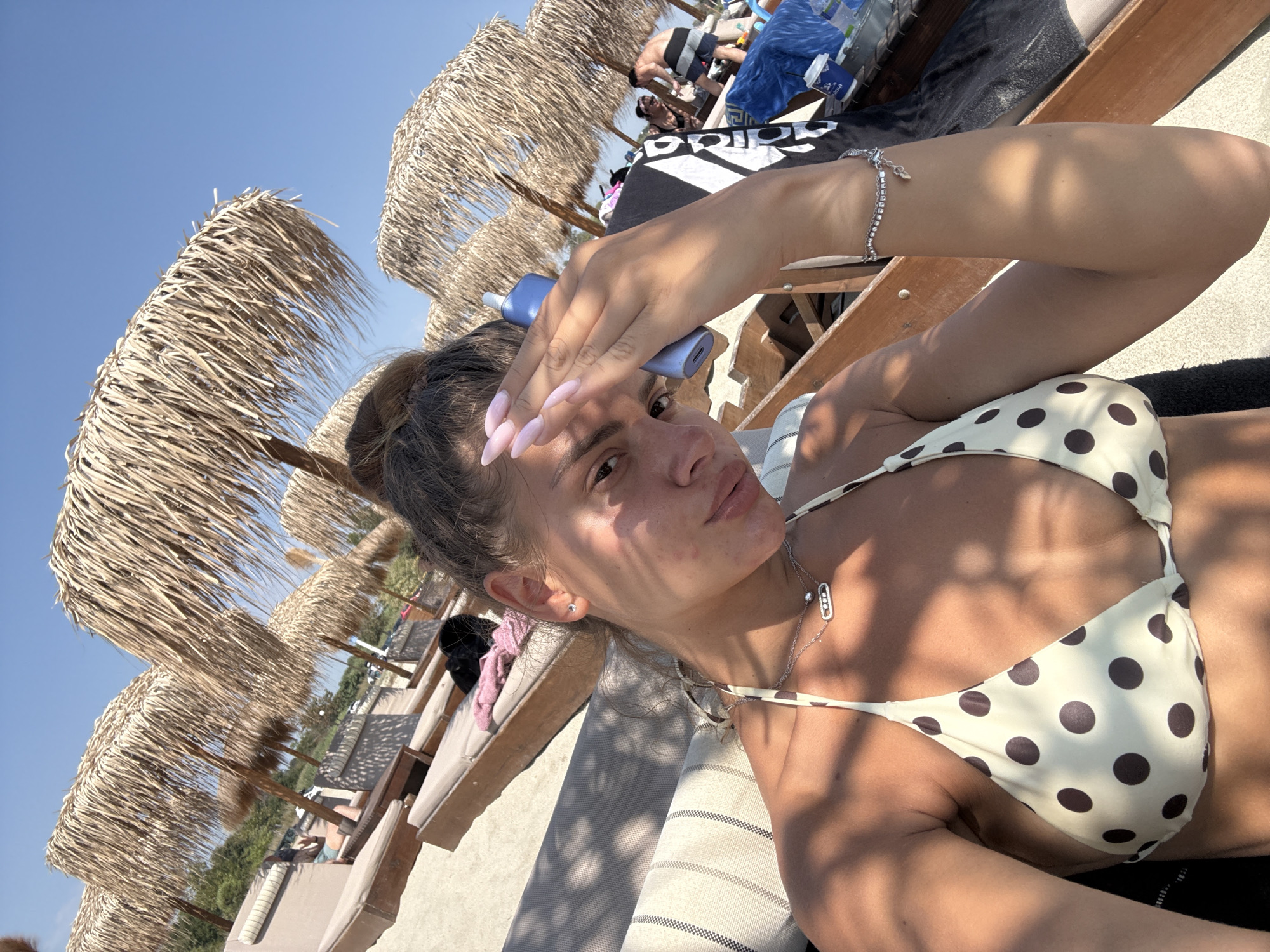 A woman is taking a selfie on a sandy beach, likely in Katerini Municipality, Greece, given the presence of straw umbrellas and lounge chairs commonly found in tourist areas. The sky is clear and blue, indicating a sunny day. The woman is wearing a polka-dot bikini top and jewelry, including a necklace and a bracelet. Her hand is raised to her forehead, possibly shielding her eyes from the sun. In the background, other beachgoers and more umbrellas are visible, along with lounge chairs and beach equipment. The overall impression is one of a relaxed vacation atmosphere.
