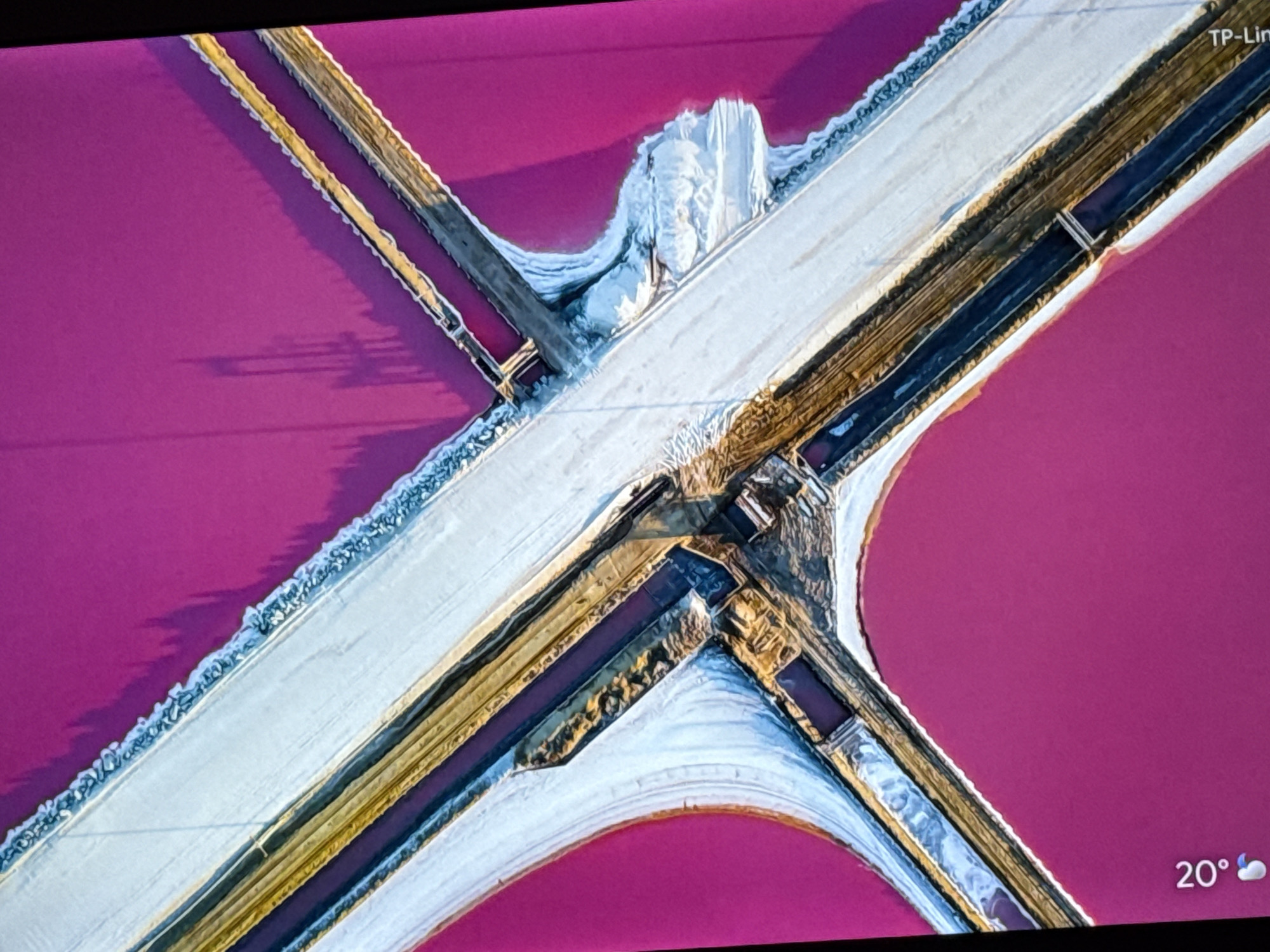 This aerial image shows a pink salt lake intersected by white and brown structures that form a large cross and a circular shape. The structures appear to be constructed from earth or salt and are lined with darker material. One section of the intersection has a buildup of white, possibly salt deposits. The lake water has a striking pink hue. The image appears to be taken from a screen, as indicated by the temperature indicator in the lower right corner reading "20°" with a moon and cloud icon, as well as "TP-Lin" in the upper right. The location is in the city of Unknown, Unknown.