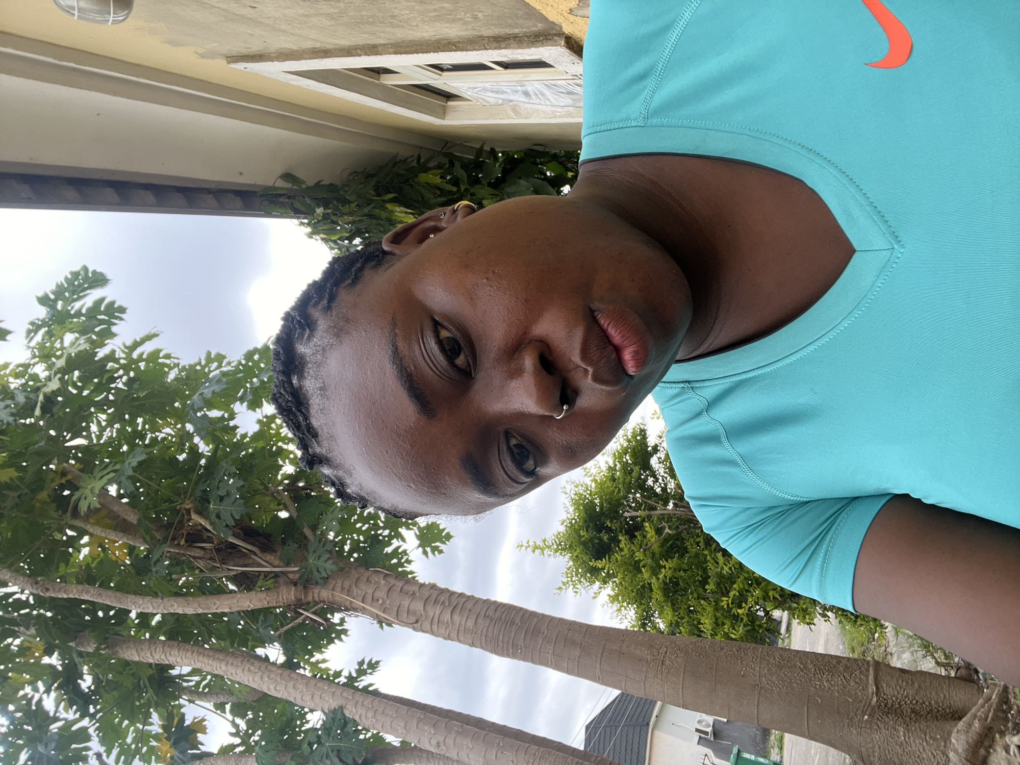 The image is a vertical, medium close-up selfie of a young Black person wearing a turquoise Nike t-shirt. Their head is tilted, and they are looking towards the camera with a neutral expression. The person has dark braided hair, a septum piercing, and an earring in their left ear.
The background suggests an outdoor setting. To the left, there's a building with a beige exterior, a window with white trim, and an outdoor light fixture. Lush green foliage from trees is visible throughout the image, with some of the trees having textured, segmented trunks. The sky is overcast, indicating a cloudy day.
There is no text visible in the image, other than the Nike logo on the t-shirt, which is an orange swoosh. The lighting suggests it is daytime, but the cloudy sky makes it difficult to determine the exact time. The overall mood is casual and relaxed, with the subject posing for a selfie. The presence of the building and the type of trees hint at a residential or possibly a public outdoor space in Abuja, Nigeria.