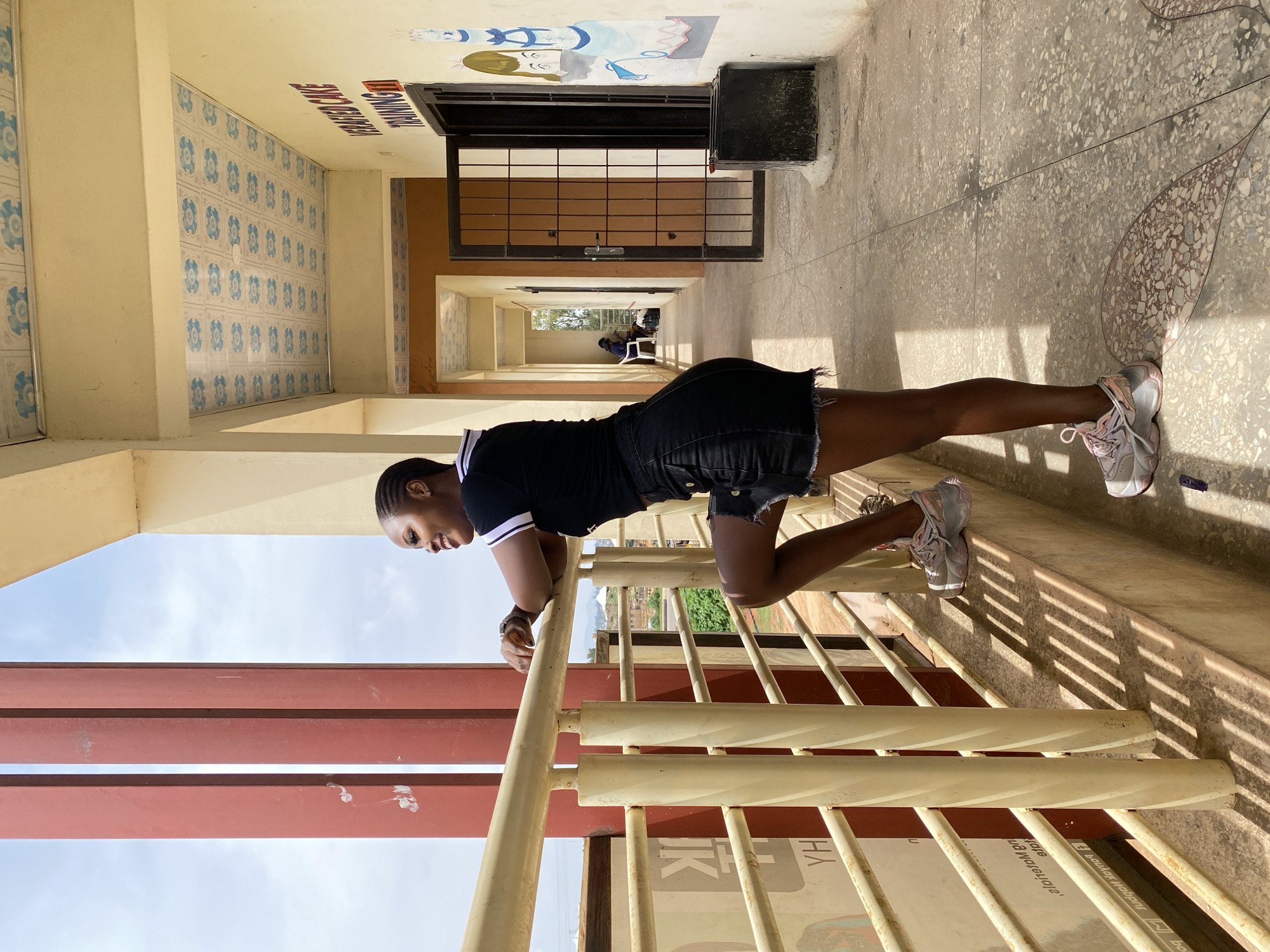 A young woman is standing on a balcony outside a building in Gura Top, Nigeria, leaning on a railing and looking to her left. She is wearing a black t-shirt with white trim and a black denim skirt. Her hair is braided and pulled back. She has on pink and white sneakers. The building has a covered walkway with a decorative tiled ceiling. A door to the right is open, revealing a darker interior. There are colorful drawings on the wall near the door, one depicting two figures. The walkway and balcony appear to be made of concrete. Shadows are cast on the ground from the railing. In the background, beyond the railing, there are some trees and structures. The sky is visible above the building, with some clouds.