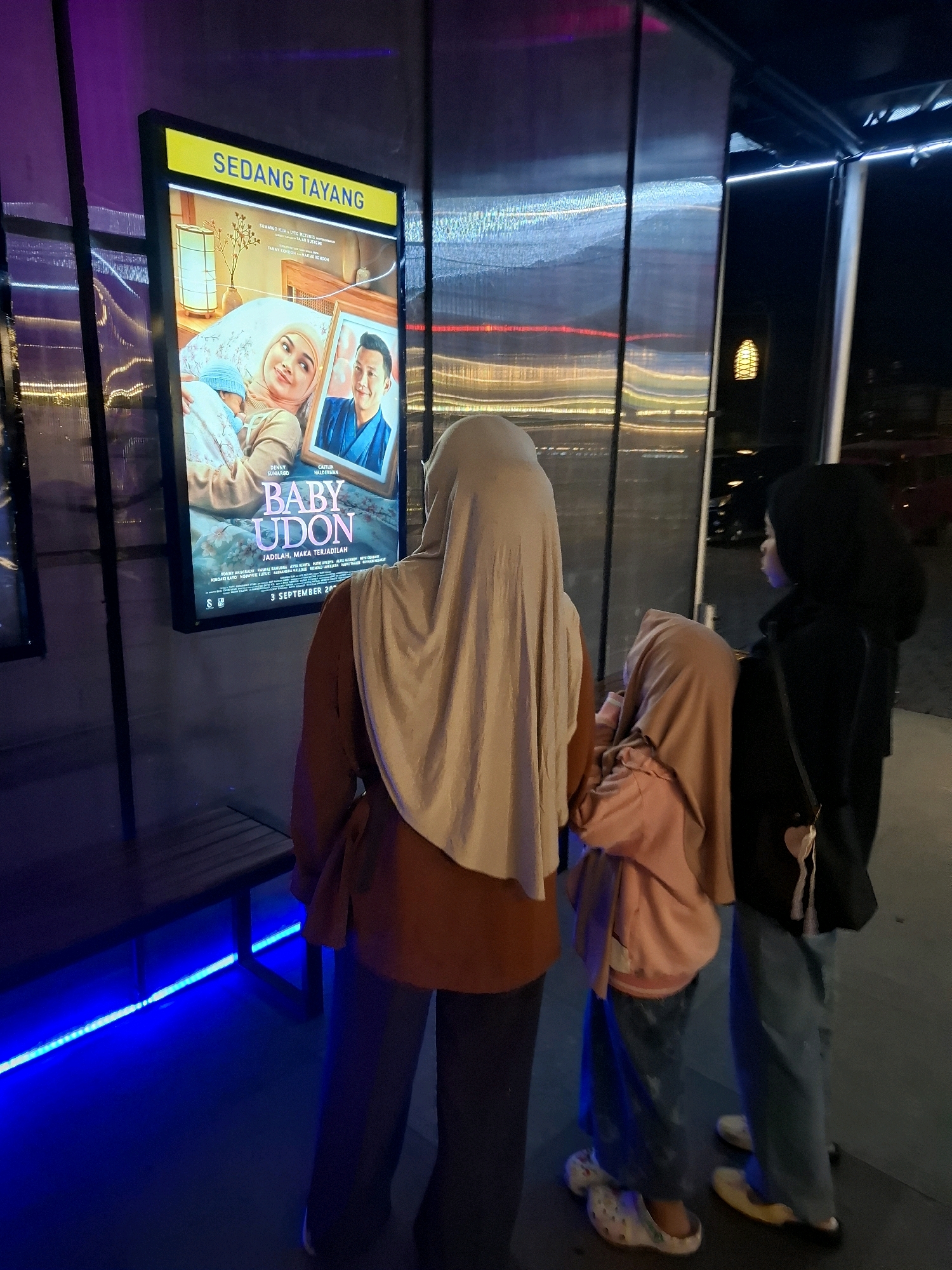 In Cianjur, Indonesia, three individuals are observed standing in front of a movie poster displayed on a brightly lit screen. The poster advertises a film titled "BABY UDON," with "SEDANG TAYANG" (Now Showing) prominently displayed at the top. The individuals, two adult women and one child, are positioned with their backs to the viewer, looking at the poster.
The individual furthest to the left, presumed to be an adult woman, wears a light beige hijab and an orange-brown jacket with dark pants. Next to her, a child, possibly a girl, is partially obscured, wearing a pink hijab and a pink hoodie over jeans. The individual on the right, also appearing to be an adult woman, is dressed in a black jacket and blue jeans.
The environment suggests an outdoor setting, possibly a cinema lobby or a public walkway. The movie poster is mounted on a dark, textured wall, illuminated by a vibrant yellow border and internal lighting. Below the poster, a dark wooden bench is visible, with a striking blue LED light strip running along its base, casting an ambient glow on the floor. In the background, a dark, reflective surface with blurred lights and architectural elements indicates a cityscape at night. The overall scene captures a moment of leisure, with the subjects engaging with the film advertisement.