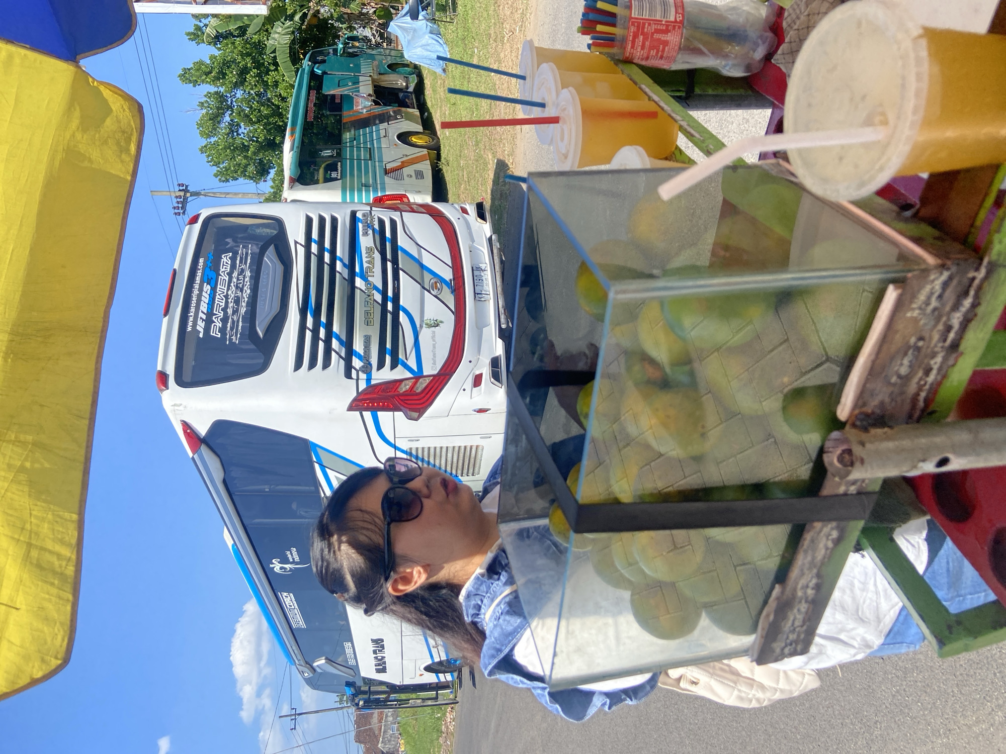 A street scene in Mangli, Indonesia, shows a person with dark hair, wearing sunglasses and a blue denim jacket, standing near a fruit stand. The stand has a glass display case filled with green fruit, with cups of orange juice with straws nearby. In the background, there are two white buses with blue stripes parked on the side of the road. The sky is blue, and the image is taken in daylight.