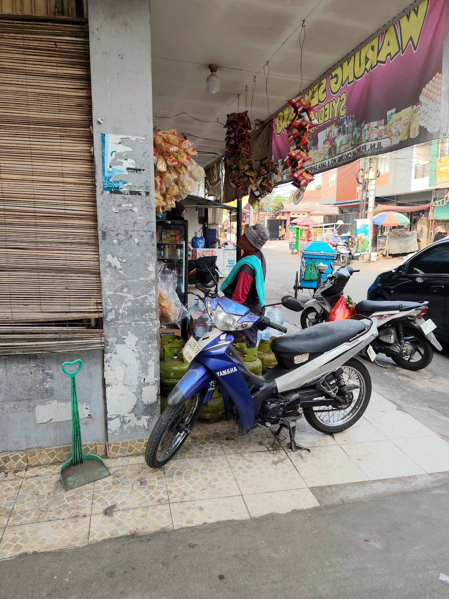 A street scene in front of a warung, or small store, in Bekasi, Indonesia. A person wearing a grey beanie, a red jacket over a teal shirt, and dark pants stands near a blue and silver Yamaha motorcycle. The motorcycle is parked on a tiled sidewalk, with several green propane tanks visible behind it. A green dustpan is leaning against a pillar. The warung has a large banner across the front advertising various products, and strings of snacks hang from the awning. Other motorcycles and a black car are visible further down the street, along with street vendors and buildings. The environment suggests a commercial street in a populated urban area.
