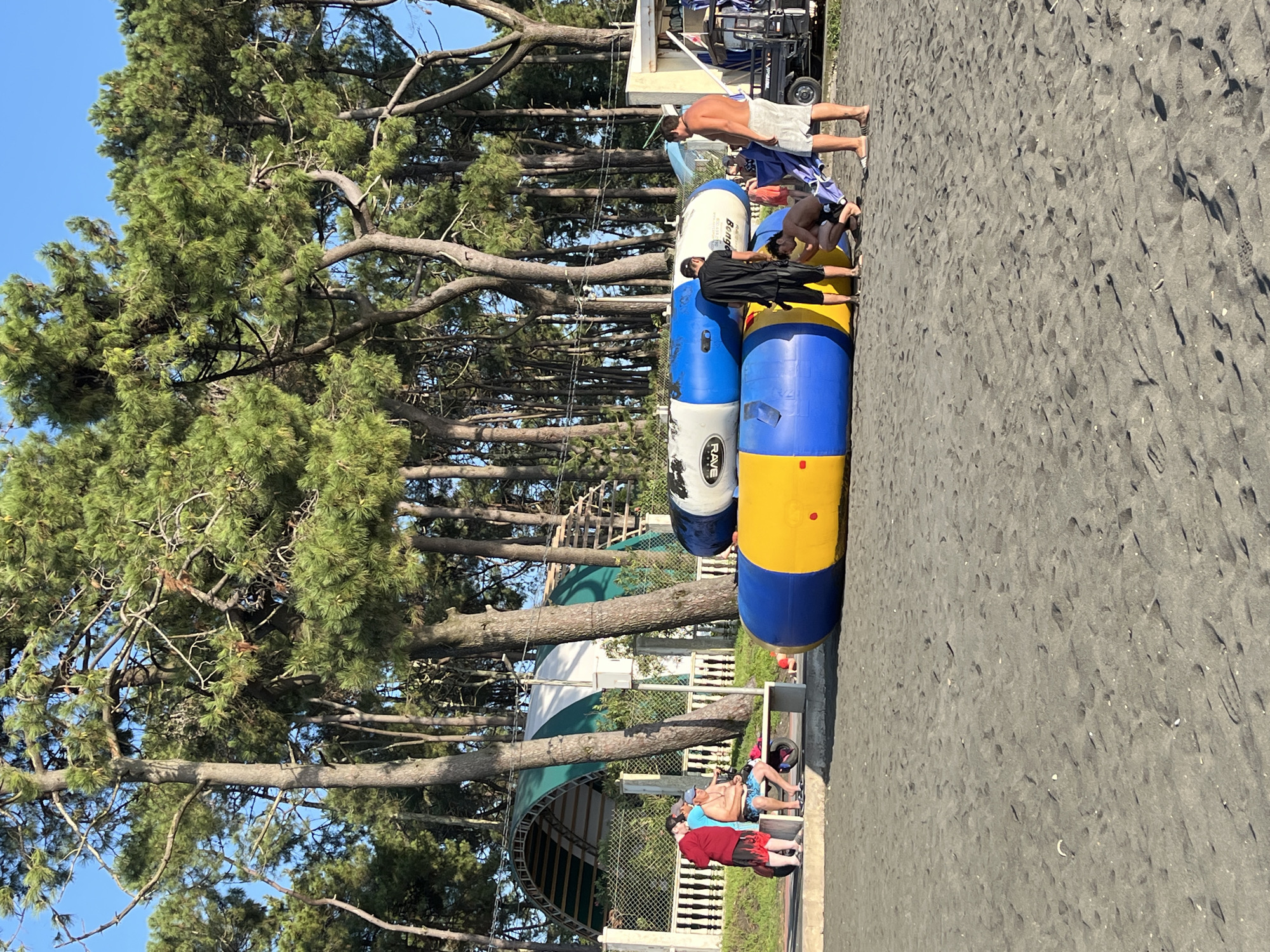 A group of people are gathered on a sandy beach near a line of pine trees in Ureki, Georgia. In the foreground, two large inflatable water toys, one blue and white and the other yellow and blue, are stacked on top of each other. Several people are standing around the inflatables, with one person in a black shirt and shorts interacting with another person in shorts who is kneeling. A man in a white shirt and shorts stands to the right, facing away from the camera. In the background, a group of people are standing and sitting on a bench near a white fence. Behind them, a large arched structure with green and white fabric is visible, and further back, more pine trees stretch towards a clear blue sky. The sand is dark and covered with small pebbles and debris, with footprints scattered across the surface.