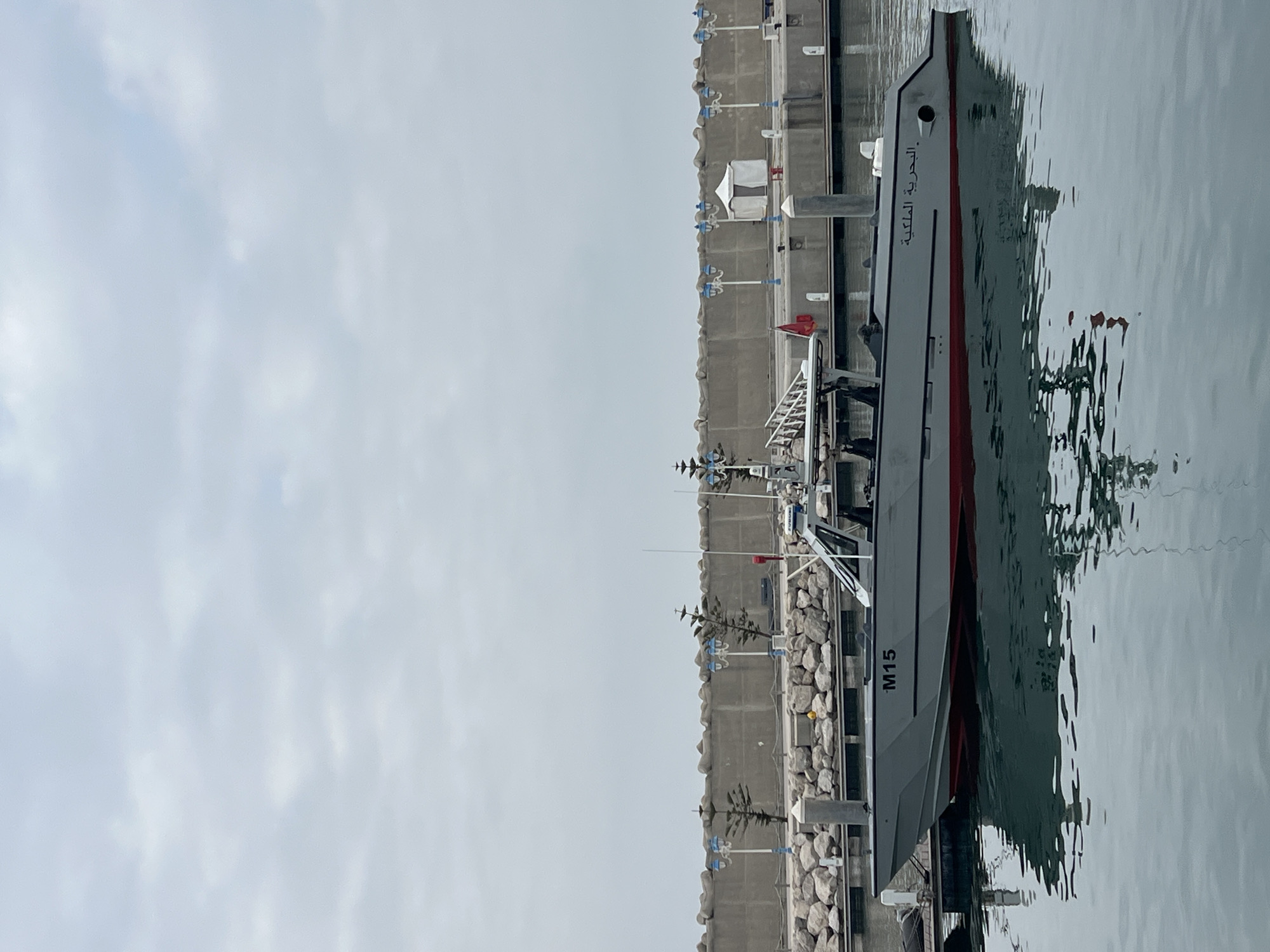 A military patrol boat is seen docked in a harbor or marina under an overcast sky. The grey vessel, featuring a red stripe along its hull, is prominently positioned on the right side of the frame. The calm water reflects the boat and the cloudy sky.
Arabic text on the bow of the boat reads "البحرية الملكية" (Al-Bahriya Al-Malakiya), which translates to "Royal Navy". Further along the hull, the designation "M15" is clearly visible. The boat is equipped with a mast, various antennas, and what appears to be radar equipment, along with a small ladder near the stern.
To the left, a sturdy concrete or stone seawall lines the harbor. Along its edge, several blue and white light fixtures or posts are installed, and large rocks form the base of the wall. A few small white tent-like structures are also present on the dock. The sky is uniformly light grey, indicating a cloudy daytime setting. No people are visible, and the scene is quiet. The presence of "Royal Navy" text confirms the vessel's military nature, and the overall setting aligns with a port in M'Diq, Morocco.