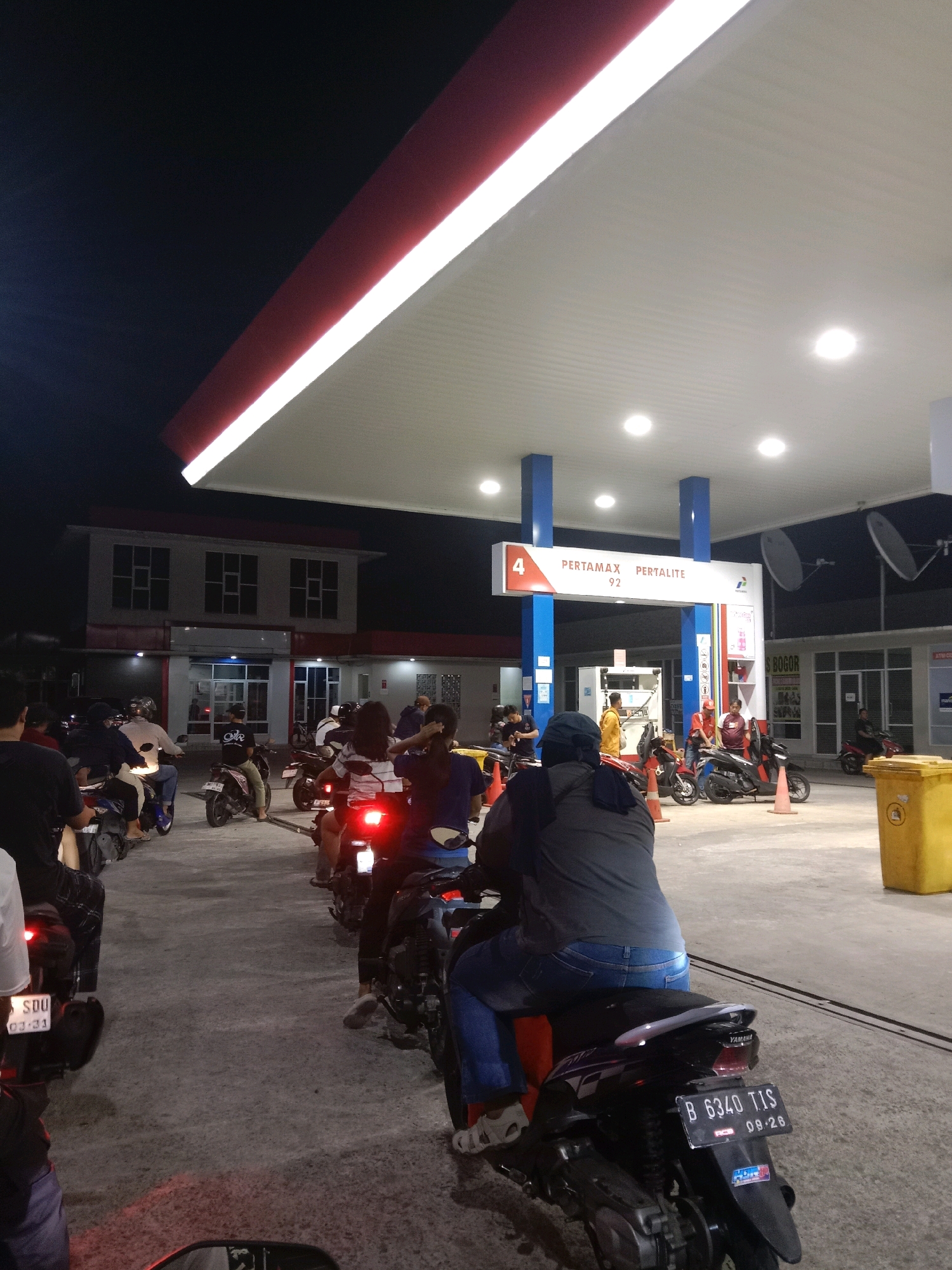 The image depicts a nighttime scene at a Pertamina gas station in the city of South Tangerang, Indonesia. A queue of motorcycles and riders is visible, waiting to refuel under the illuminated canopy of the gas station. The canopy features red and white trim and is supported by blue pillars. Signs indicate "PERTAMAX PERTALITE 92." In the background, a two-story building with multiple windows and red trim is present. Several people are on motorcycles, some wearing helmets, and one person in the foreground is wearing a gray shirt and blue jeans. A yellow trash bin is situated to the right of the frame. The ground is paved concrete, and the lighting suggests it is after dark.
