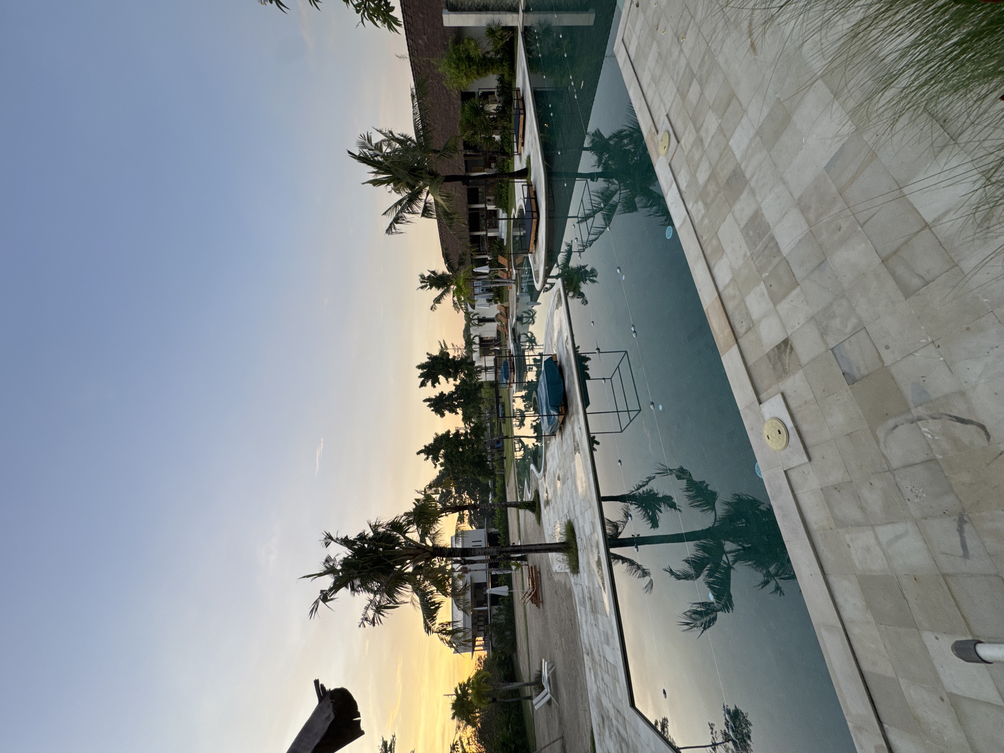 The image depicts a pool at sunset in Kuta, Indonesia, reflecting palm trees and nearby buildings. The sky transitions from blue to yellow. Palm trees line the pool's edge. The pool's tiles are light-colored. There is no evidence of people.