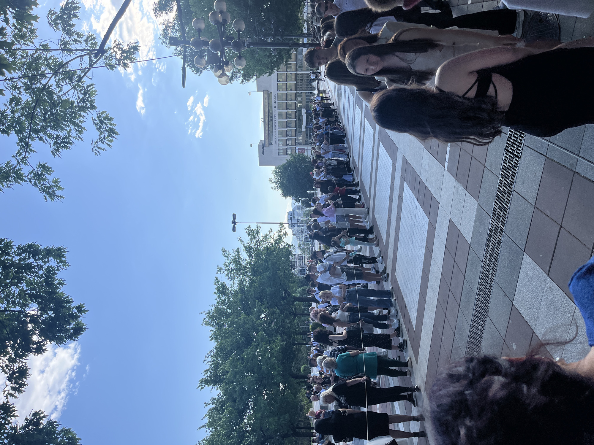 A large group of people stand hand-in-hand in a line along a pedestrian walkway. The background shows a city environment with trees, buildings, and a sky with some clouds. The location is in Blagoevgrad, Bulgaria, specifically in front of the American University in Bulgaria.