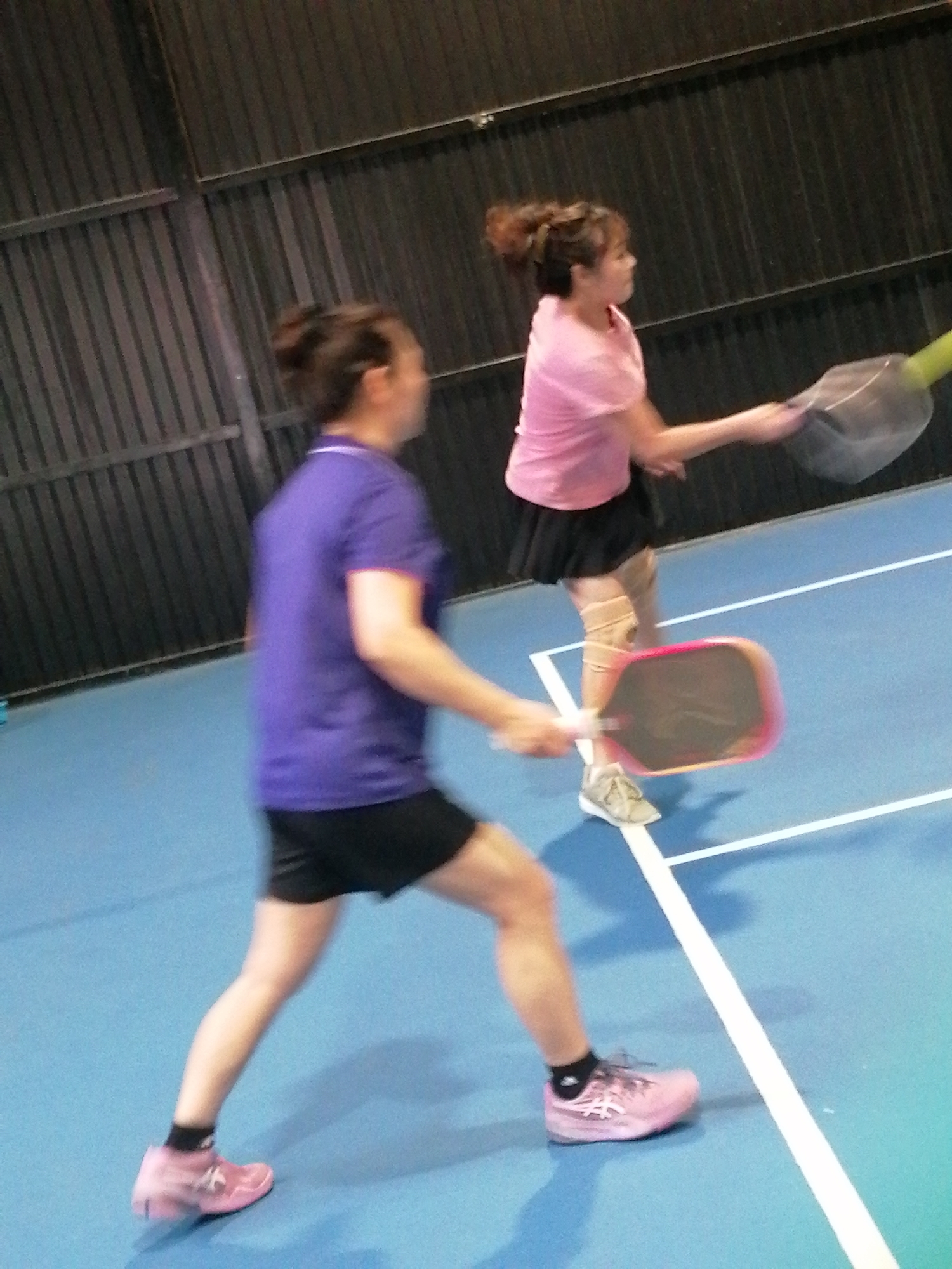 Two women are playing pickleball indoors on a blue court with white lines. The woman in the foreground is wearing a purple shirt and black shorts, holding a pink pickleball paddle. She is in motion, with her legs bent and her left foot forward. The woman in the background is wearing a pink shirt and a black skirt, also holding a pickleball paddle. She is facing the net and appears to be in the process of hitting the ball. The court has a dark, corrugated metal wall in the background. The location is identified as Cam Ly - Da Lat, Vietnam.