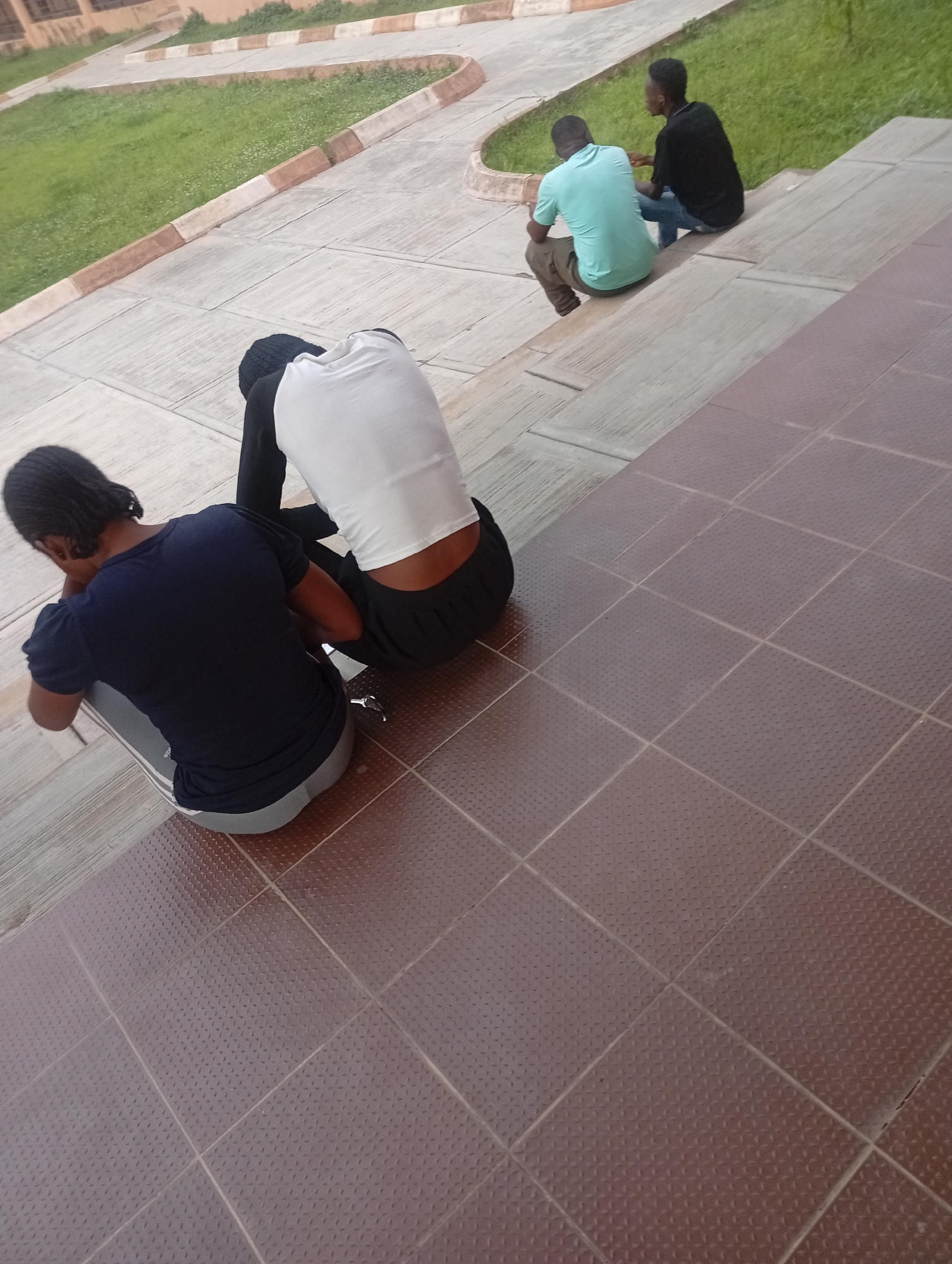 The image shows a group of young men sitting on outdoor steps in Yelwa, Nigeria. In the foreground, two individuals are seated with their backs to the camera. One is wearing a dark blue t-shirt and grey pants, and the other is wearing a white t-shirt and black pants. Further up the steps, two other young men are visible, one in a teal t-shirt and khaki pants, and the other in a black t-shirt and blue jeans. They are all facing away from the camera, with the latter two appearing to be in conversation. The steps are made of concrete and have a tiled surface in the foreground. Behind the steps, there is a grassy area and a building visible in the distance.