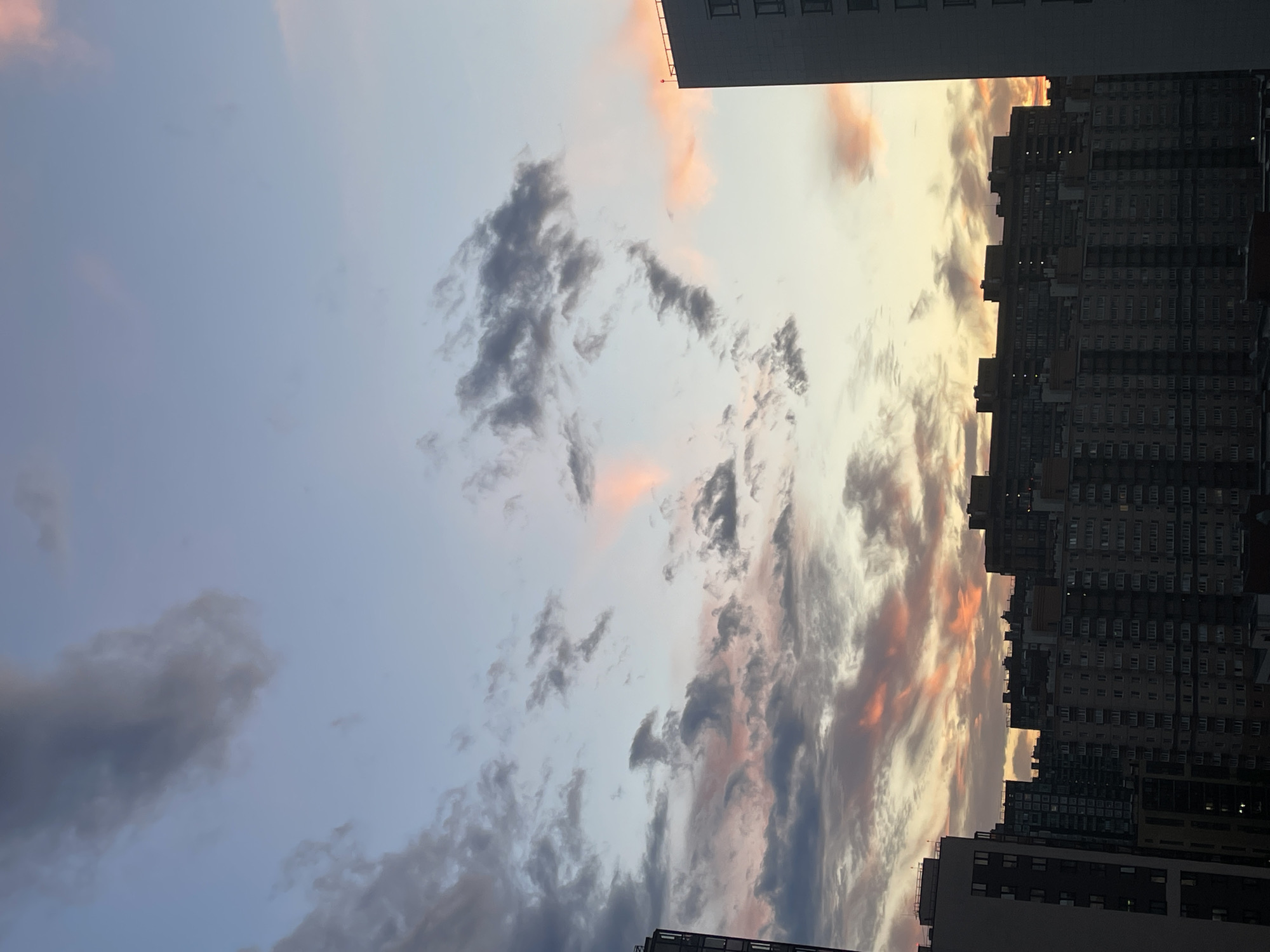 The image shows a city skyline at dusk. Tall buildings, some with visible windows and balconies, dominate the right side of the frame. The sky is filled with dramatic clouds, painted with hues of pink, orange, and gray, suggesting the setting sun. The overall mood is serene and atmospheric, capturing the quiet beauty of a city transitioning from day to night. The orientation of the photo is vertical.