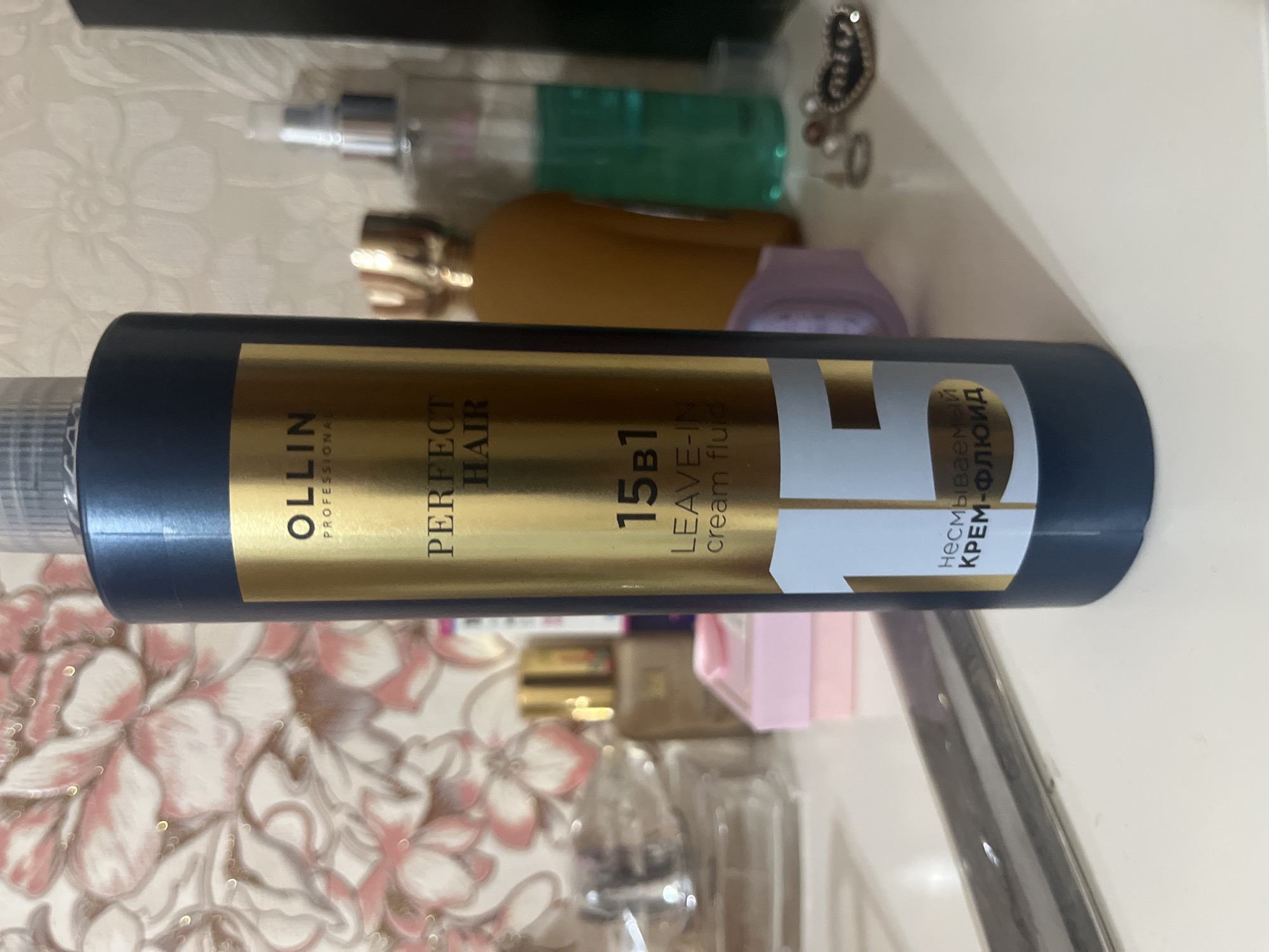 The image shows a dark blue bottle of "OLLIN PROFESSIONAL PERFECT HAIR 15IN1 LEAVE-IN CREAM FLUID" standing on a white surface, likely a table or shelf. The bottle has a gold label with black text. Behind the bottle, a wall with a floral pattern is visible. To the right of the bottle, several other objects are present, including a light brown bottle, a clear bottle with green liquid, some jewelry, and what appears to be a small pink box. The scene is indoors, and there is no visible indication of the city of Almaty, Kazakhstan, or any specific urban environment. The lighting suggests an interior setting, possibly a room with artificial or natural light sources. No people or actions are depicted in the image.