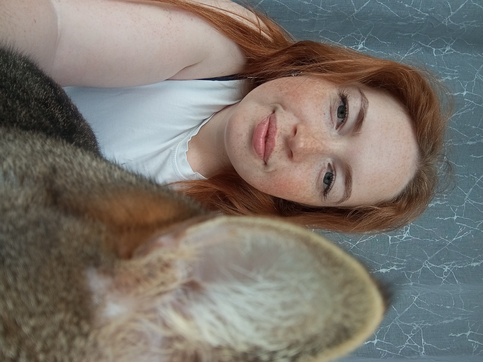 A woman with red hair and freckles is lying on a patterned gray surface, possibly a bed or blanket. She is wearing a white t-shirt. A cat, primarily brown and gray, is positioned in the foreground, obscuring part of the woman's body. The image was taken indoors. The specific location of the image is not possible to determine from the image alone.