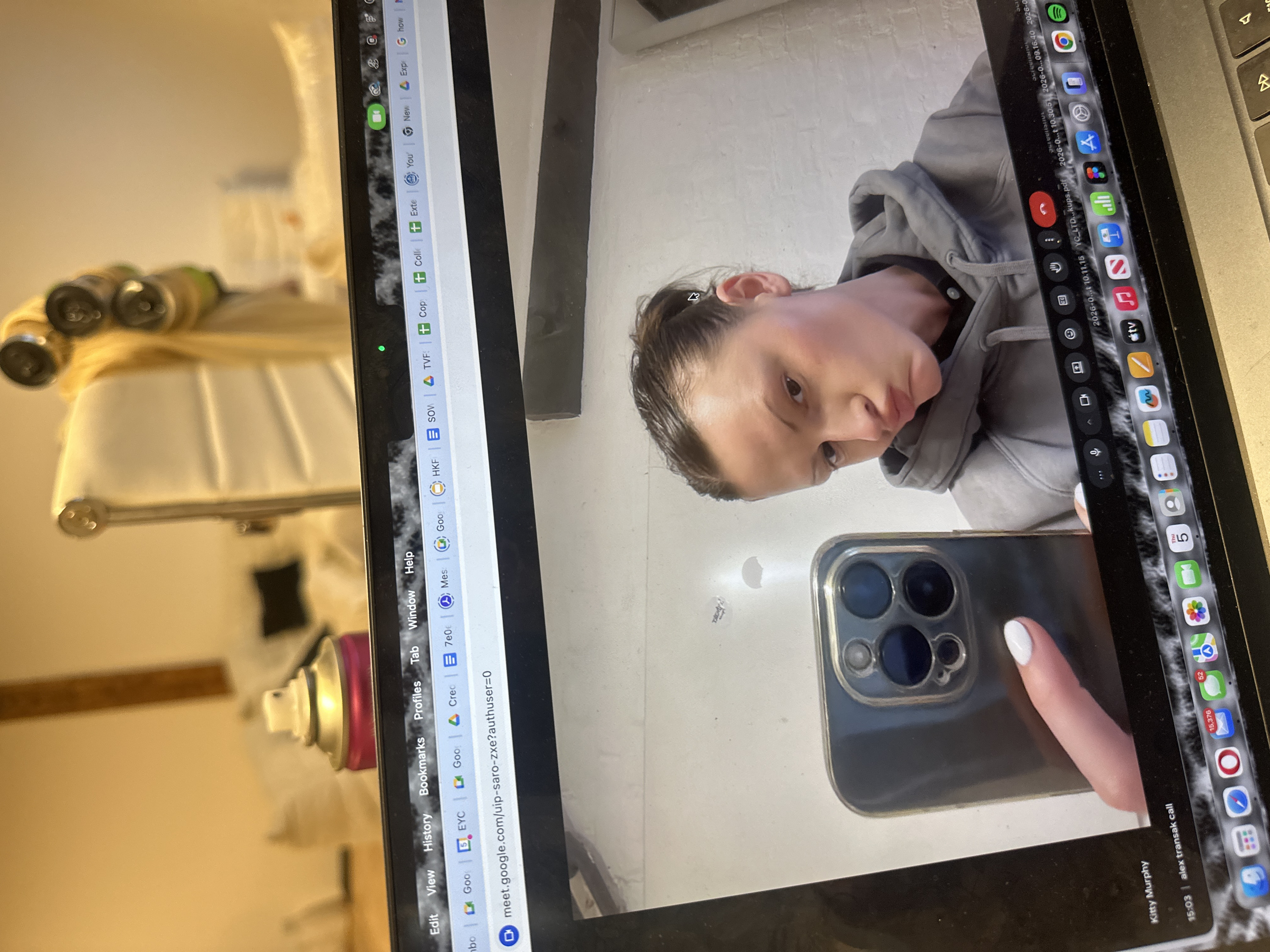 The image displays a photograph taken of a laptop screen, oriented horizontally, with various items visible in the surrounding physical environment.
On the laptop screen, a video call or a mirrored image shows a person (likely female) taking a selfie with a smartphone. The person is looking downwards and slightly to their right, with dark hair pulled back. They are wearing a grey hooded sweatshirt with a dark collar visible underneath. Their right hand, with a finger sporting white nail polish, holds a dark-colored smartphone featuring a prominent multi-lens camera module. The background visible on the phone's screen or behind the person on the laptop screen is a light-colored, textured wall.
The laptop's interface is clearly visible around the video call content.
* **Top browser bar**: Numerous tabs are open, including icons for Google, Google Meet, YouTube, Spotify, and various generic browser icons. A Google Meet URL (`meet.google.com/uip-saro-zxe?authuser=0`) is readable in one of the tabs.
* **Right-hand dock/taskbar**: Displays a row of application icons. Visible apps include FaceTime (green camera icon), Settings (gears), App Store, Photos, Mail (showing "15,376" unread emails), Safari, Messages, Calendar, Music, Apple TV, and several document icons with dates like "2026-0...". A red notification bubble with "52" is present over one icon.
* **Bottom left of the screen**: Text reads "Kitty Murphy" followed by "15:03" and "alex transak call", indicating the time and participants of a call or recent communication.
To the left of the laptop, several objects are visible in the physical environment, appearing slightly out of focus. These include:
* Multiple hair rollers, which are silver/metallic with some green accents, appear to be holding blonde or light-colored hair.
* A blurry white object with vertical ridges, possibly a piece of furniture like a headboard or chair.
* A pink and silver spray can, likely hair spray, oriented horizontally with its nozzle pointing towards the screen.
The scene suggests an afternoon setting (15:03) where someone is either on a video call or using their laptop to display their phone's camera feed, possibly while styling their hair. The numerous open tabs and notifications indicate active computer usage.