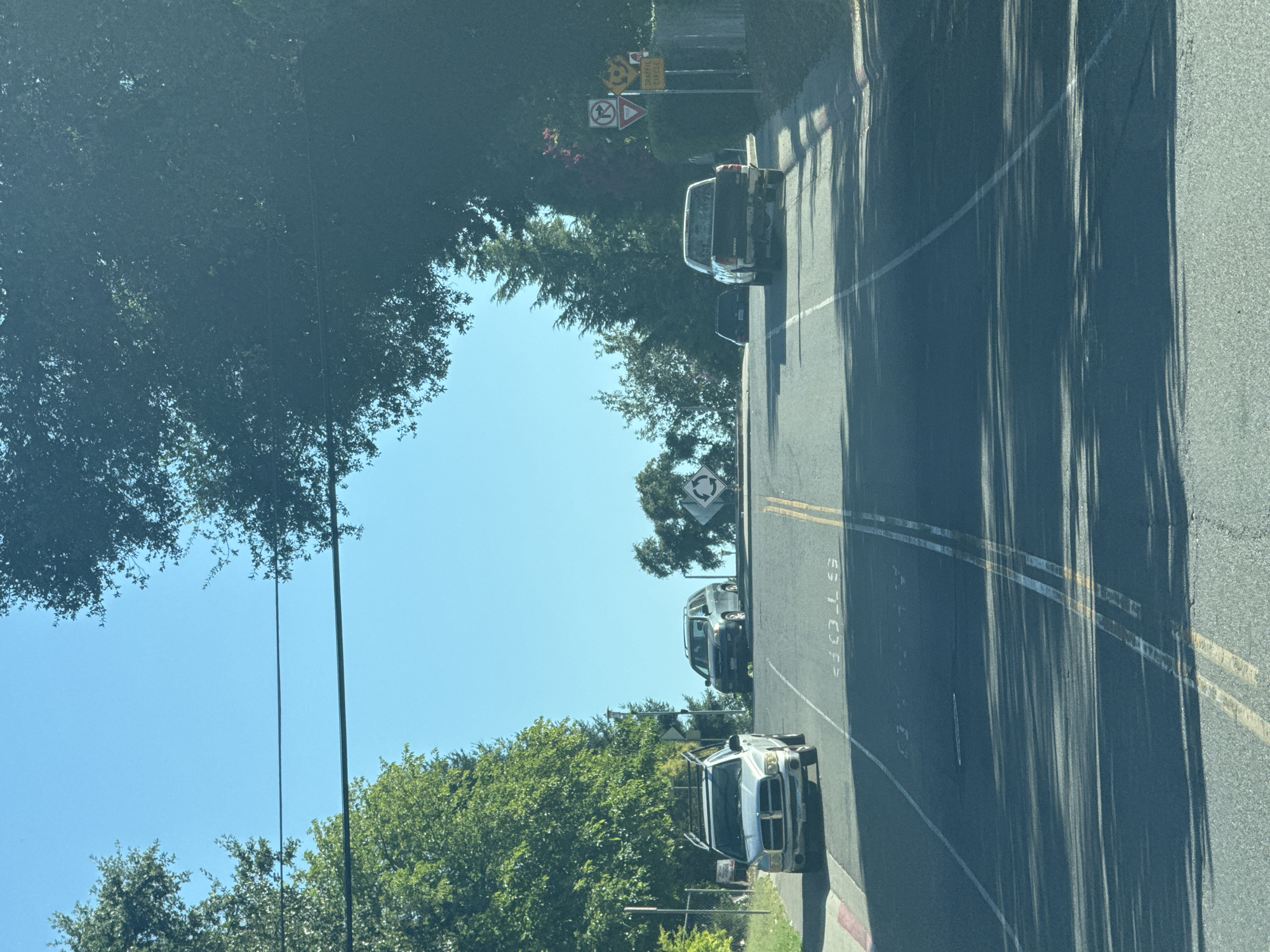The image depicts a street scene in Citrus Heights, California, USA, on a sunny day. The view is from inside a vehicle, looking up a gently sloping road lined with trees and residential properties. Several cars are visible on the road: a silver pickup truck in the left lane, a dark-colored SUV in the right lane, and another pickup truck in the right lane further up the hill. Road markings, including a "STOP" sign painted on the asphalt, are present. Traffic signs indicating a "TRAFFIC CIRCLE" and a "NO LEFT TURN" are visible on the right side of the road. The sky is clear blue, and the sun casts shadows from the trees onto the road.