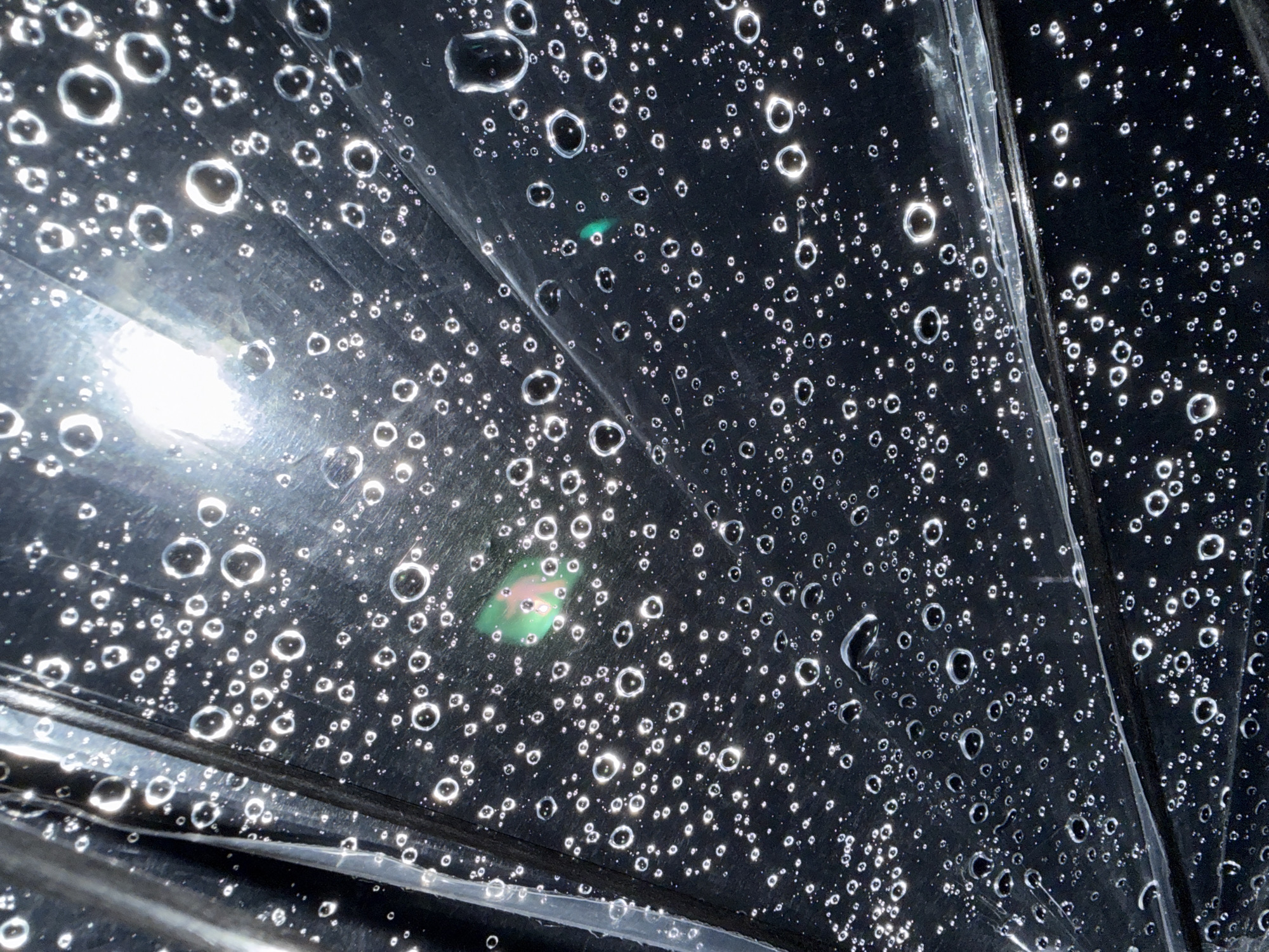 The image is an extreme close-up of a black, reflective surface covered in numerous small water droplets. The droplets vary in size, with some appearing as perfect circles and others as irregular shapes. The surface appears to be made of a smooth, possibly plastic material, with a textured, ribbed section visible in the upper left and diagonally across the lower portion of the frame. A bright light source is reflected in the upper center of the image, causing lens flare and highlighting the individual droplets. There are also faint green and pink iridescent glints visible on the surface in some areas, possibly due to the way light is refracting through the water. There are no people or animals present in the image. The image does not contain any identifiable landmarks or features that would allow for localization within Yachiyo, Japan.